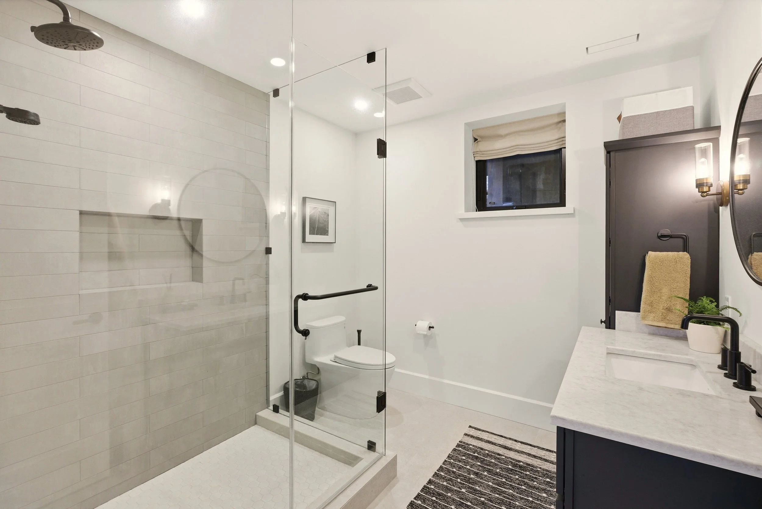 Modern bathroom with glass shower enclosure, white toilet, window with beige shade, black cabinet, white marble countertop with black fixtures, small potted plant, and black and white striped rug.