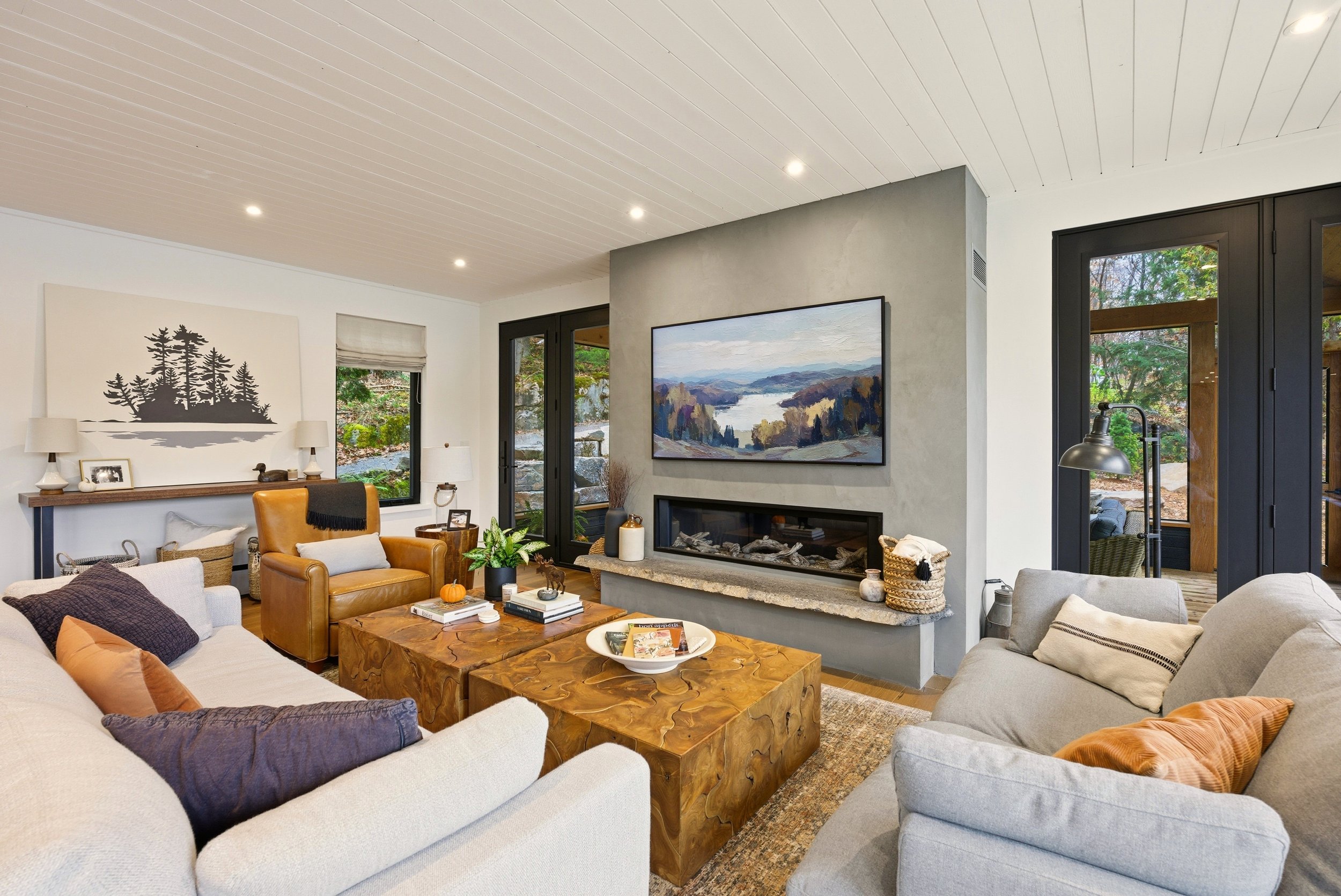 Living room with beige sofas, a brown leather armchair, a wooden coffee table, a fireplace, and large windows showing greenery outside.