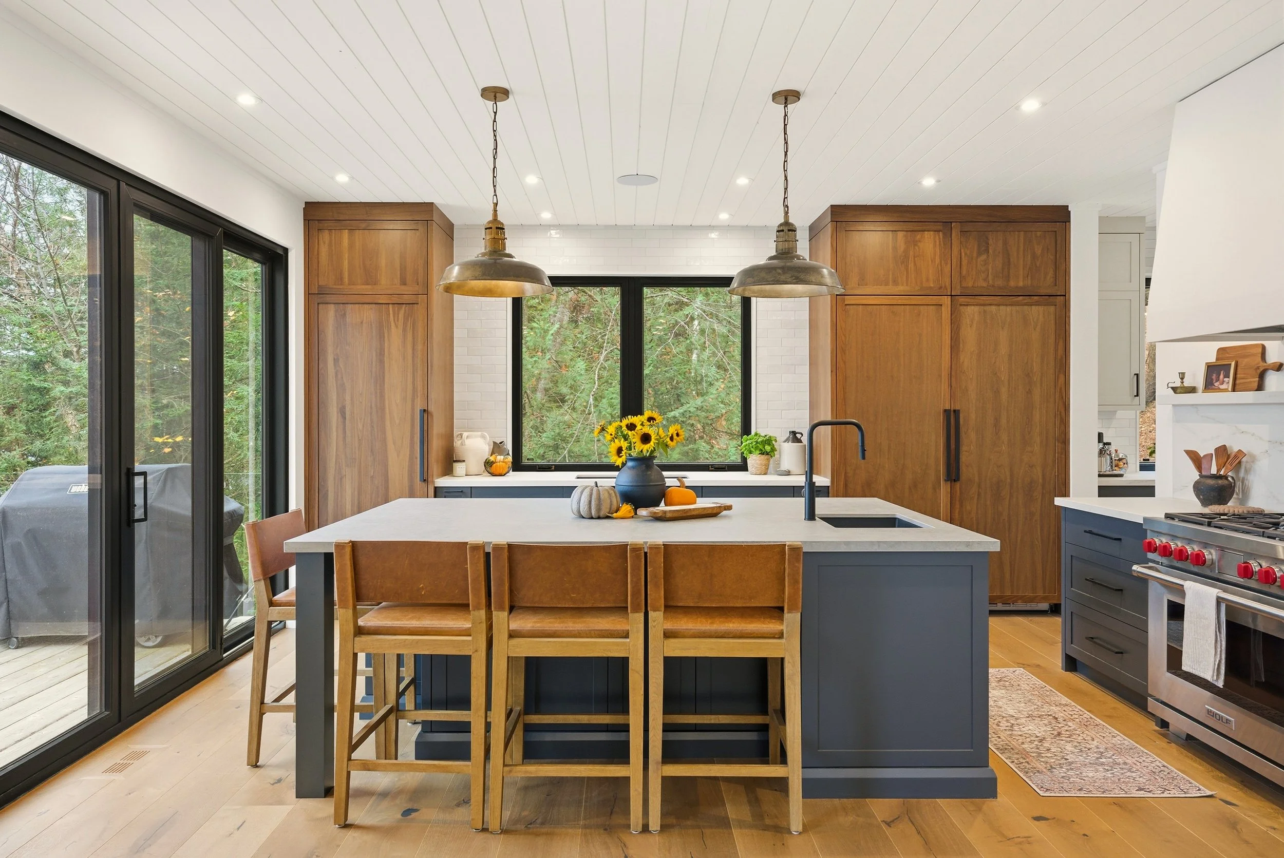 Modern kitchen with wooden cabinets, an island with bar stools, large window, sliding glass door, and outdoor grill.