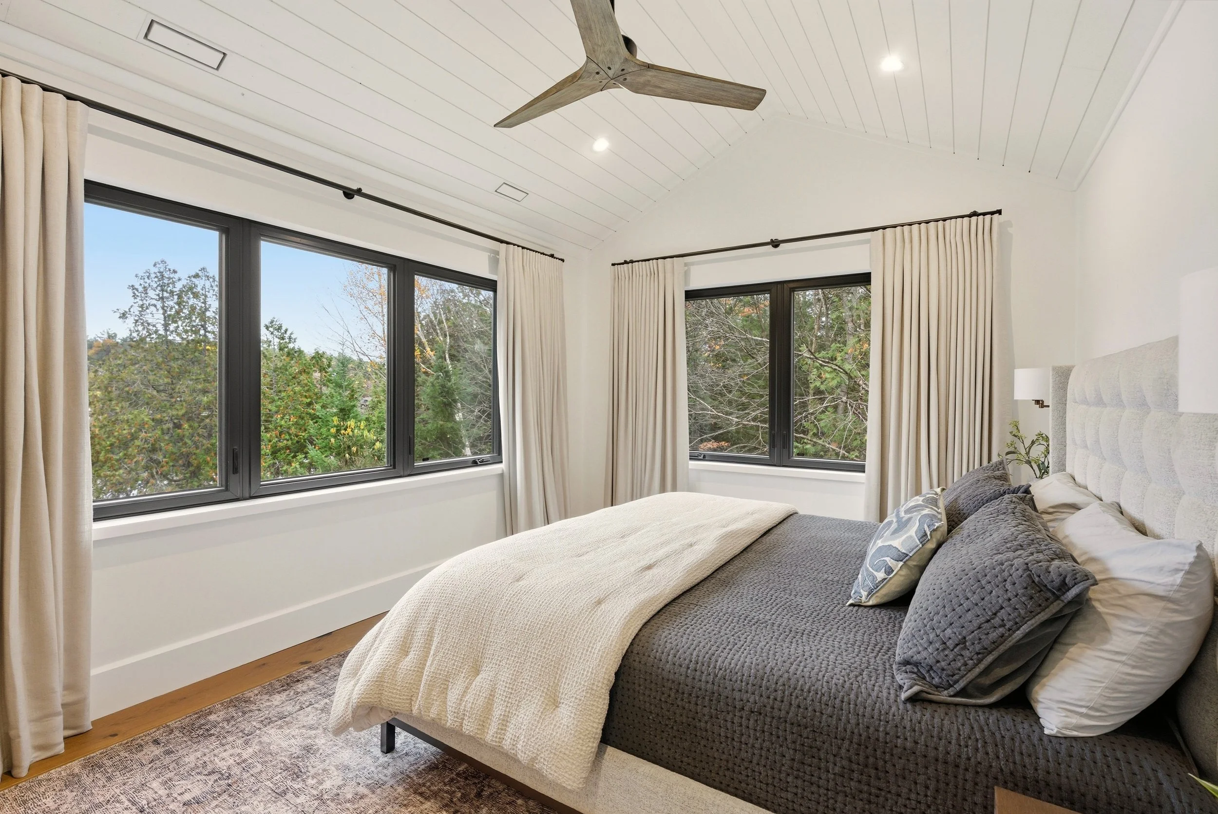 Bedroom with large windows, beige curtains, a gray bed with multiple pillows, a beige blanket, and a wooden ceiling fan.