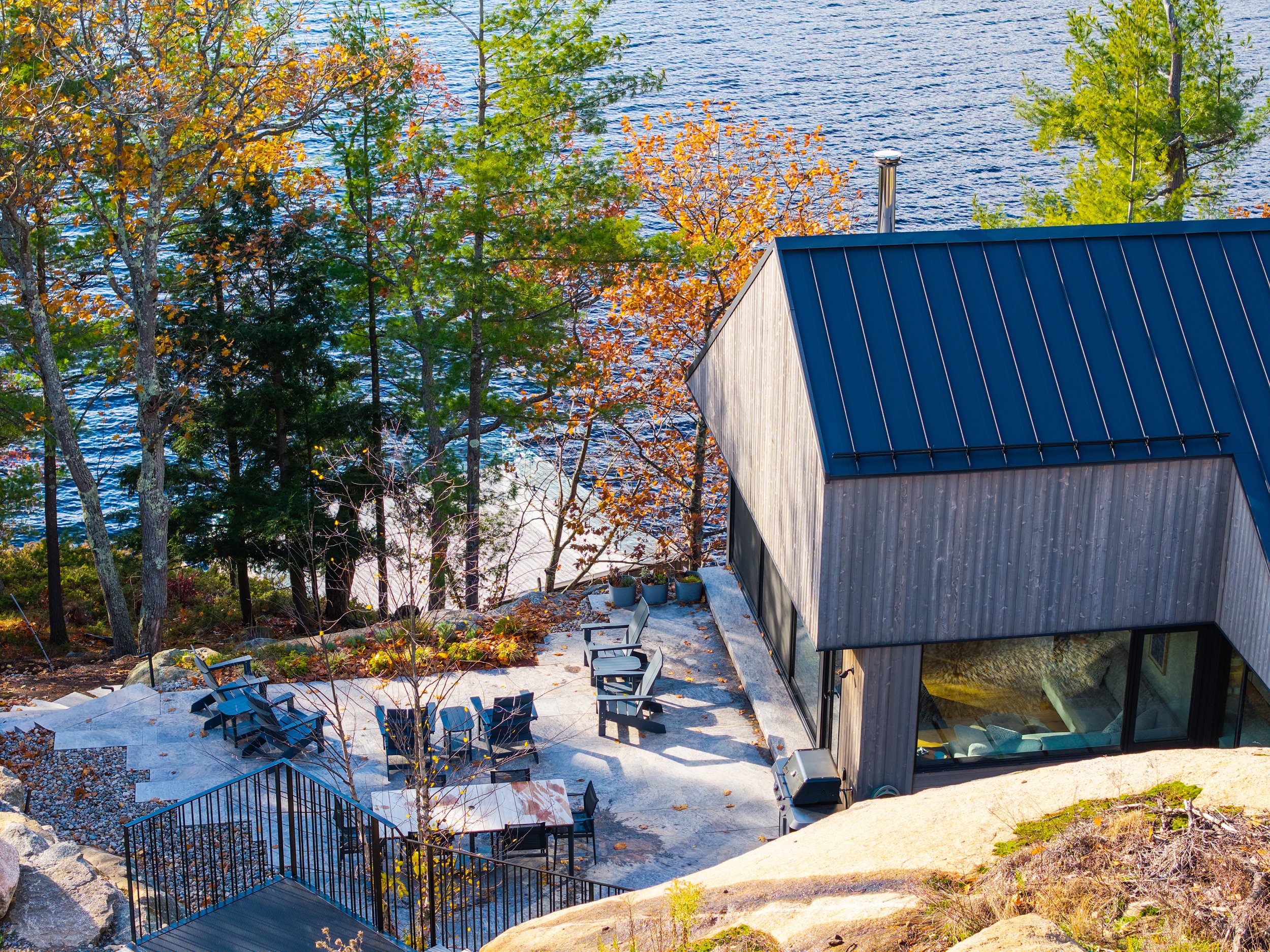 A modern house with a dark blue metal roof, situated beside a body of water, surrounded by autumn trees, with a patio area featuring chairs and potted plants.