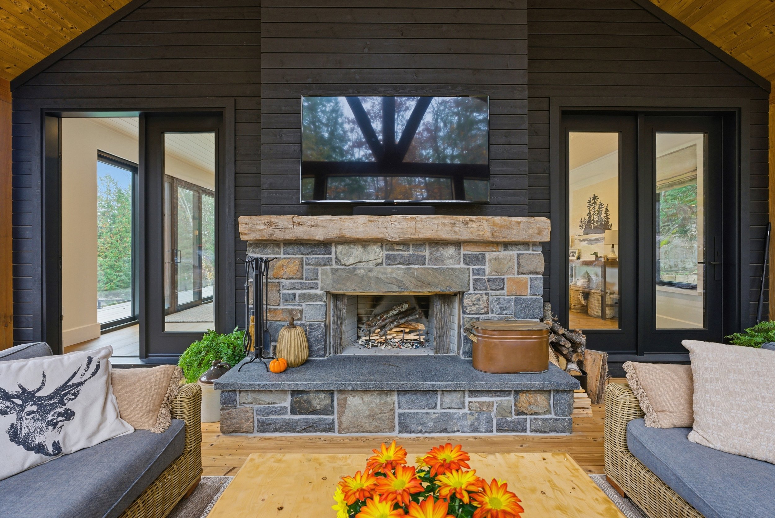 Living room with a stone fireplace in Muskoka, built by Conklin's Construction, a flat-screen TV above it, and sliding glass doors on either side leading to outdoor spaces.