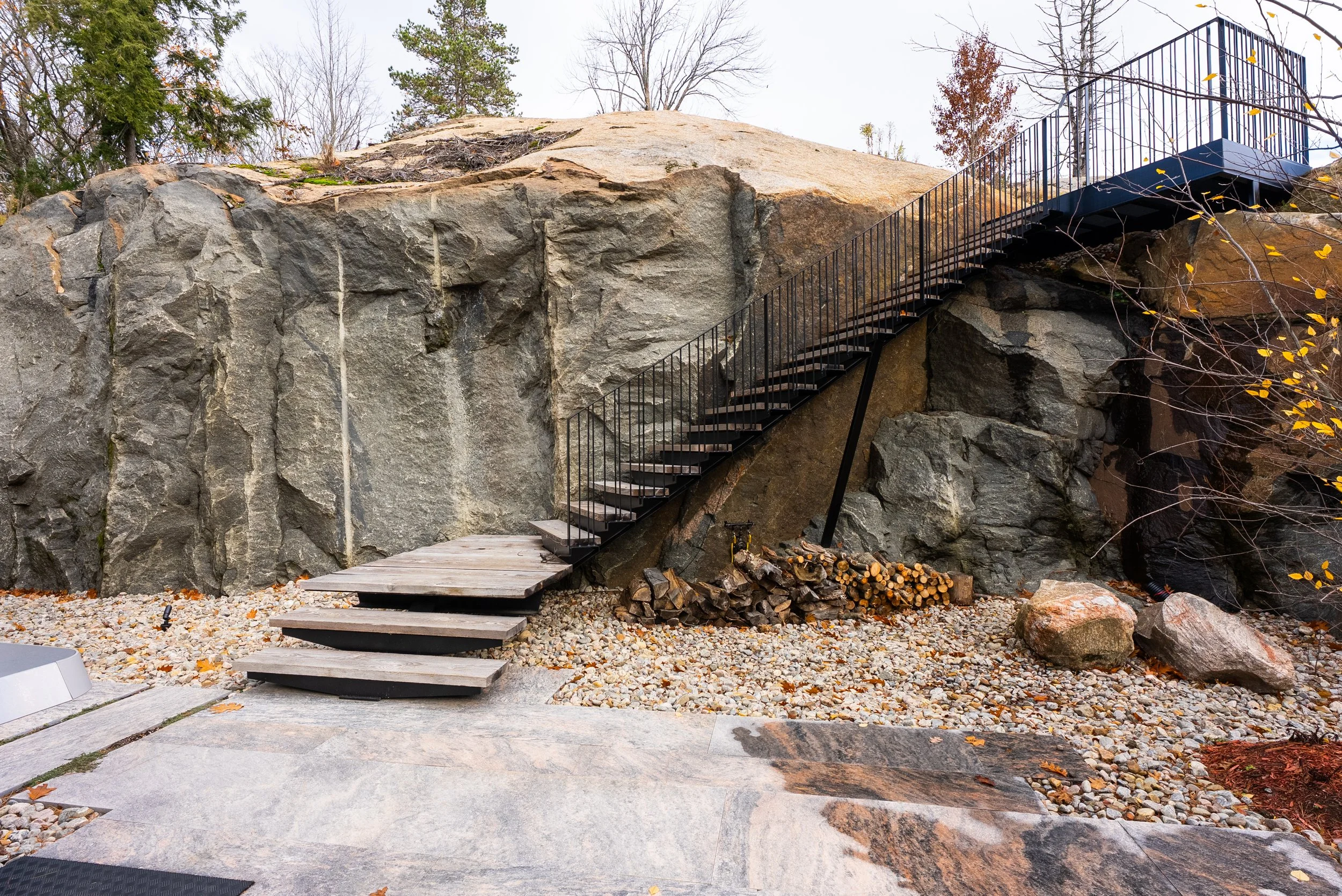 Outdoor scene with a rock wall and a modern metal staircase leading upward to a platform, surrounded by trees and rocks.