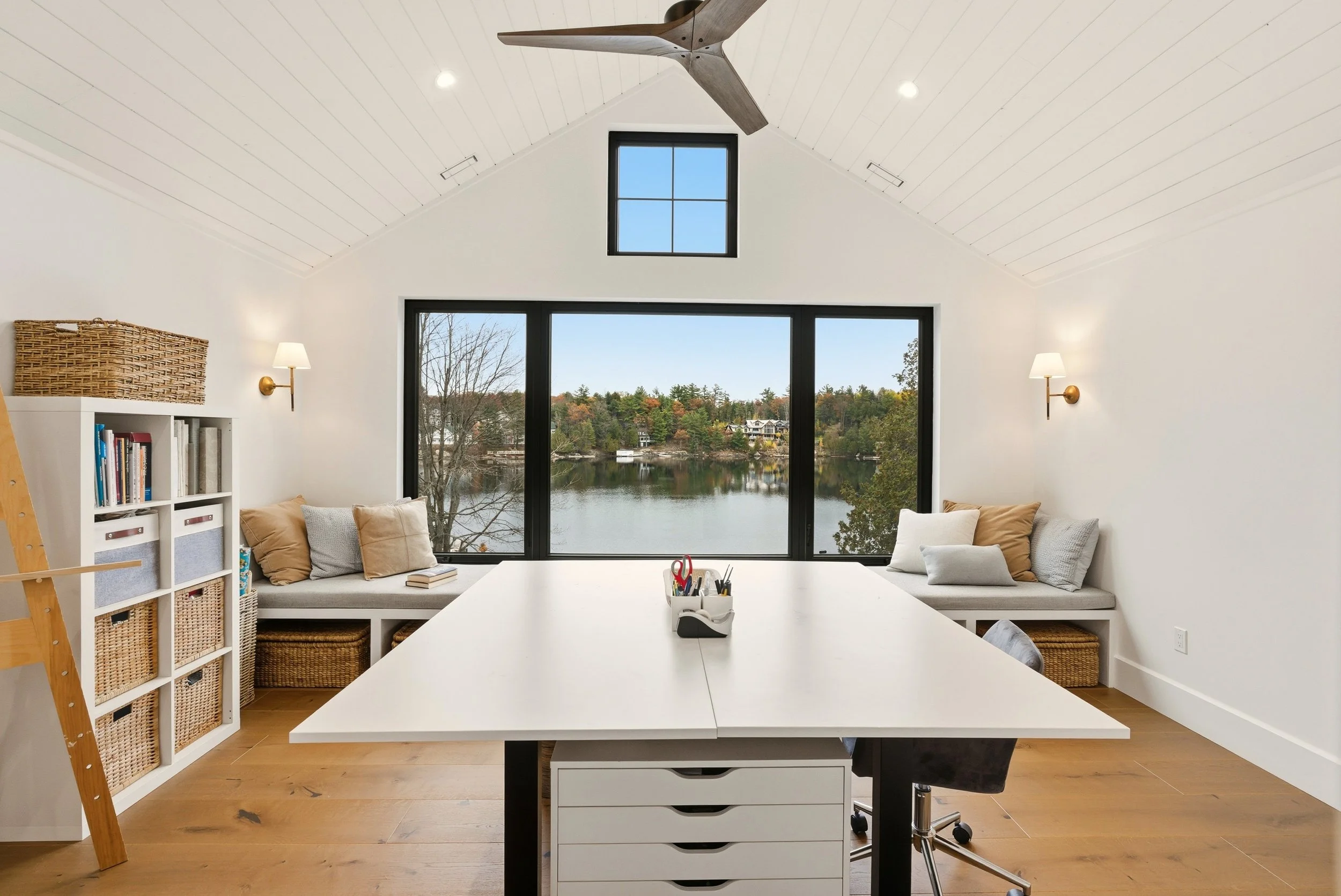 A bright home office with a large window overlooking a lake, white walls, a white desk, built-in window seats with cushions, and a view of trees and houses outside.