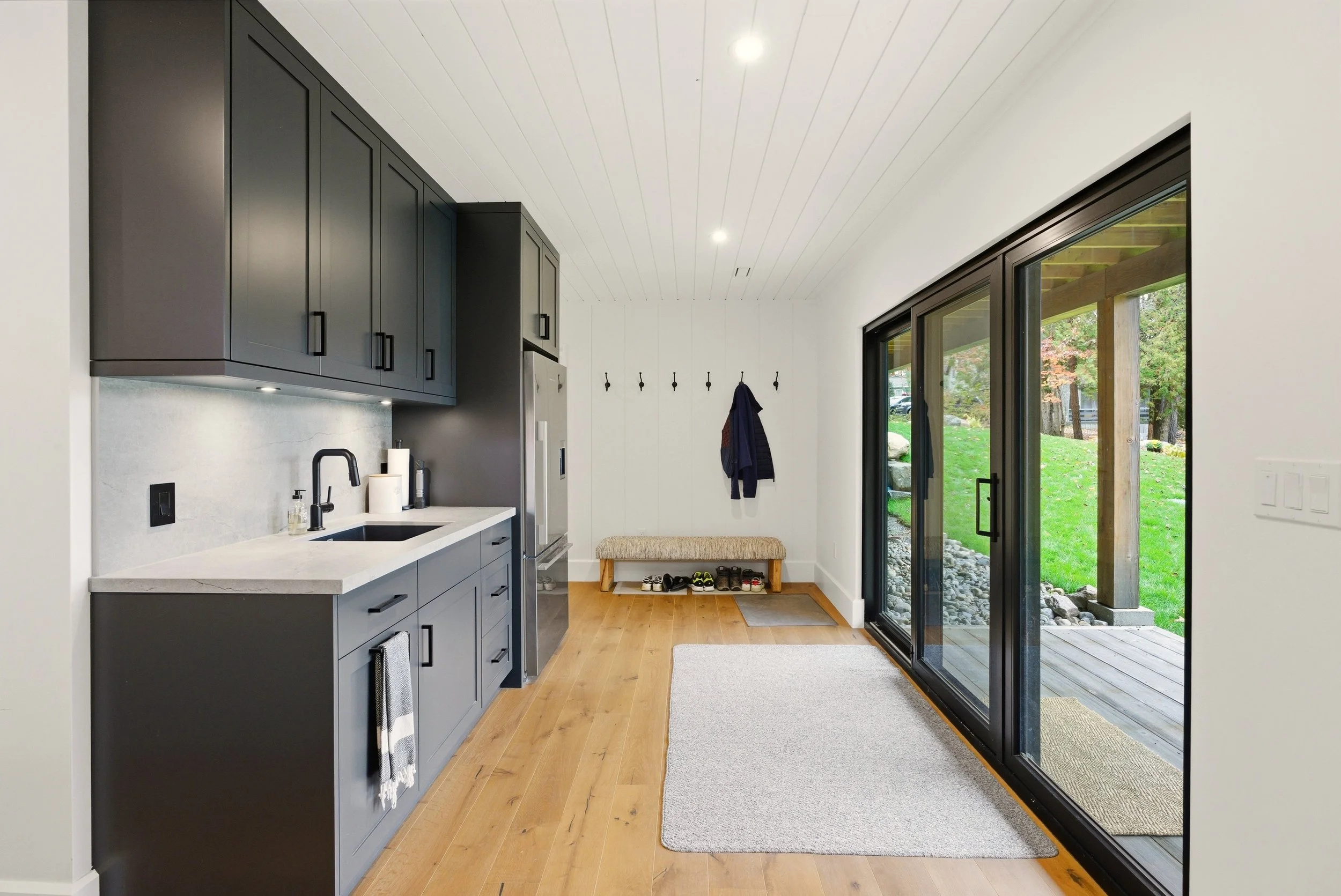 Interior view of a modern kitchen with black cabinets, white countertops, a stainless steel refrigerator, and a large sliding glass door leading to a deck and green outdoor space.