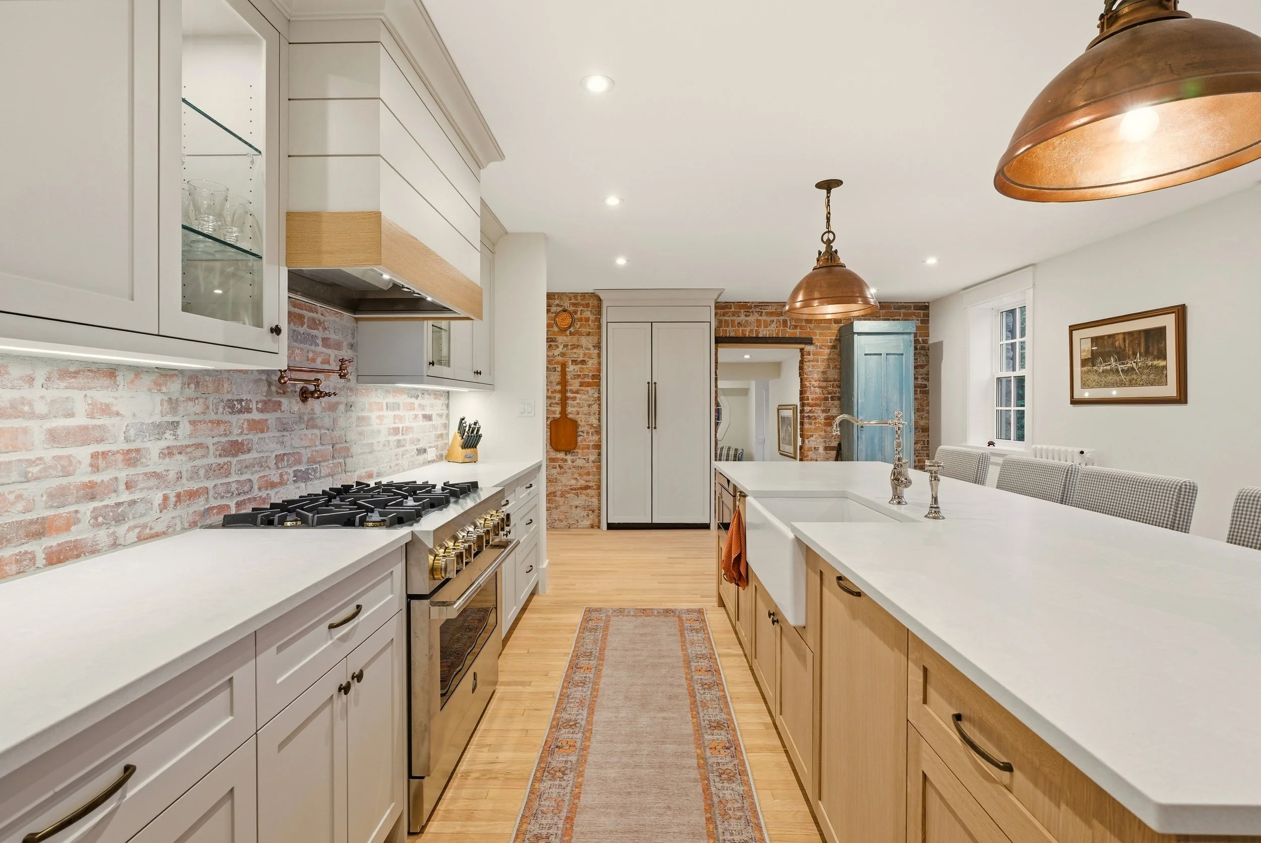 A modern kitchen with white cabinets, brick backsplash, a central island with a farmhouse sink, and pendant lights. There is a built-in oven, a row of barstools, and a hardwood floor.