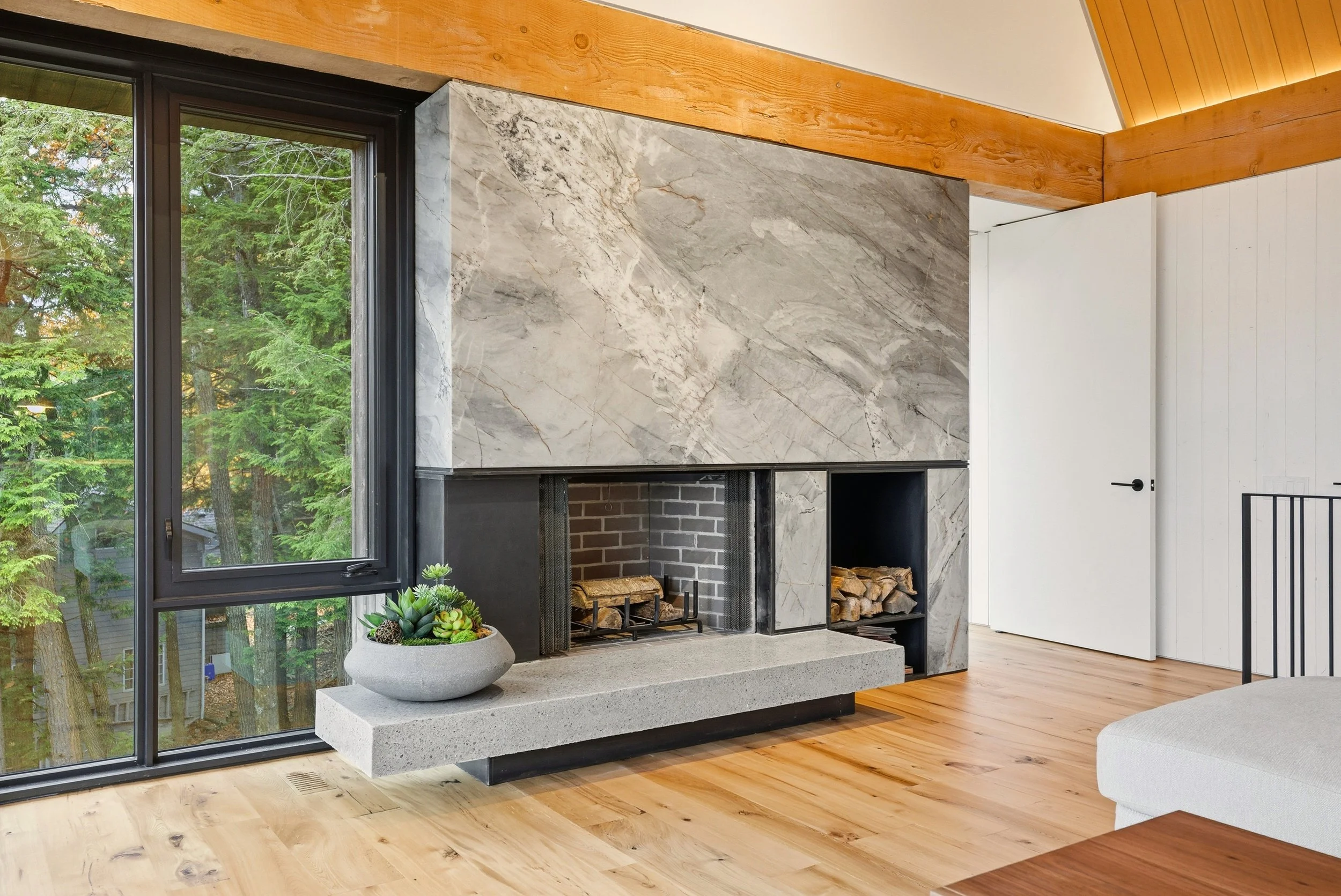 Interior view of a living room featuring a large fireplace with marble surround, a glass sliding door to the left showing green trees outside, a small gray planter with succulents on the concrete hearth, hardwood floor, white paneled wall, and an ope