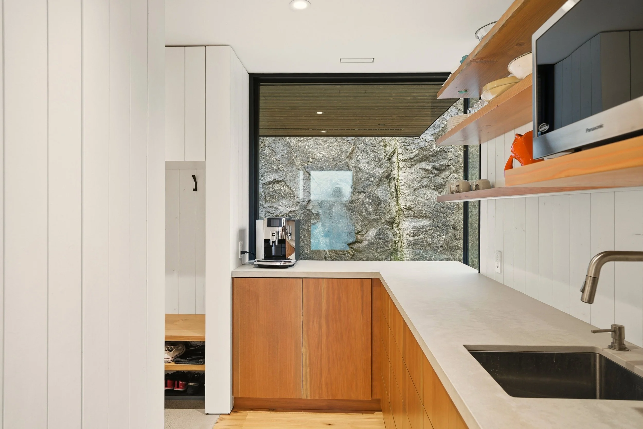 Modern kitchen with white wood-paneled walls, wooden cabinets, a coffee machine on the counter, and a large window overlooking a rock wall.