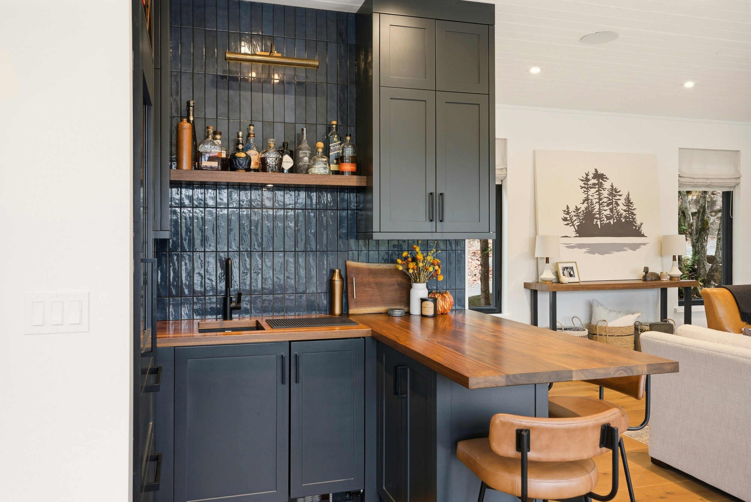 Kitchen with dark blue cabinets, wooden countertop, black tiled backsplash, and open shelving with bottles. Small kitchen island with tan leather stool. Adjacent living area with artwork of trees and a white couch.