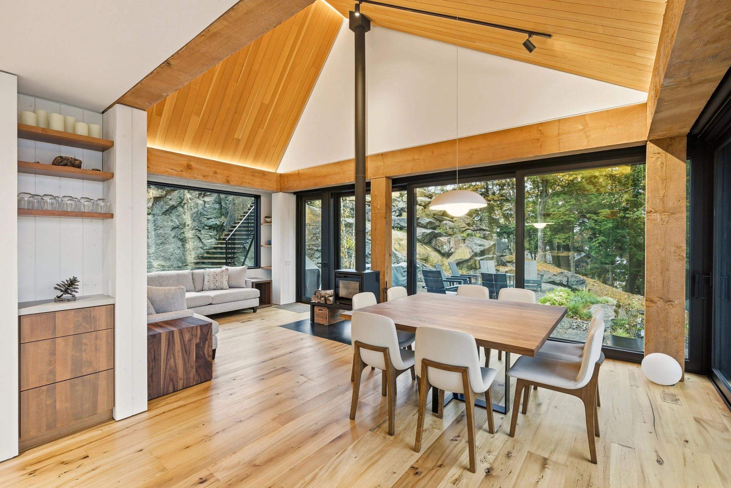 Open-concept living and dining room with wood-paneled ceiling, large sliding glass doors, and a view of rocks and greenery outside. Features a wooden dining table with eight white chairs, a modern fireplace, and a sitting area with a sofa and armchai