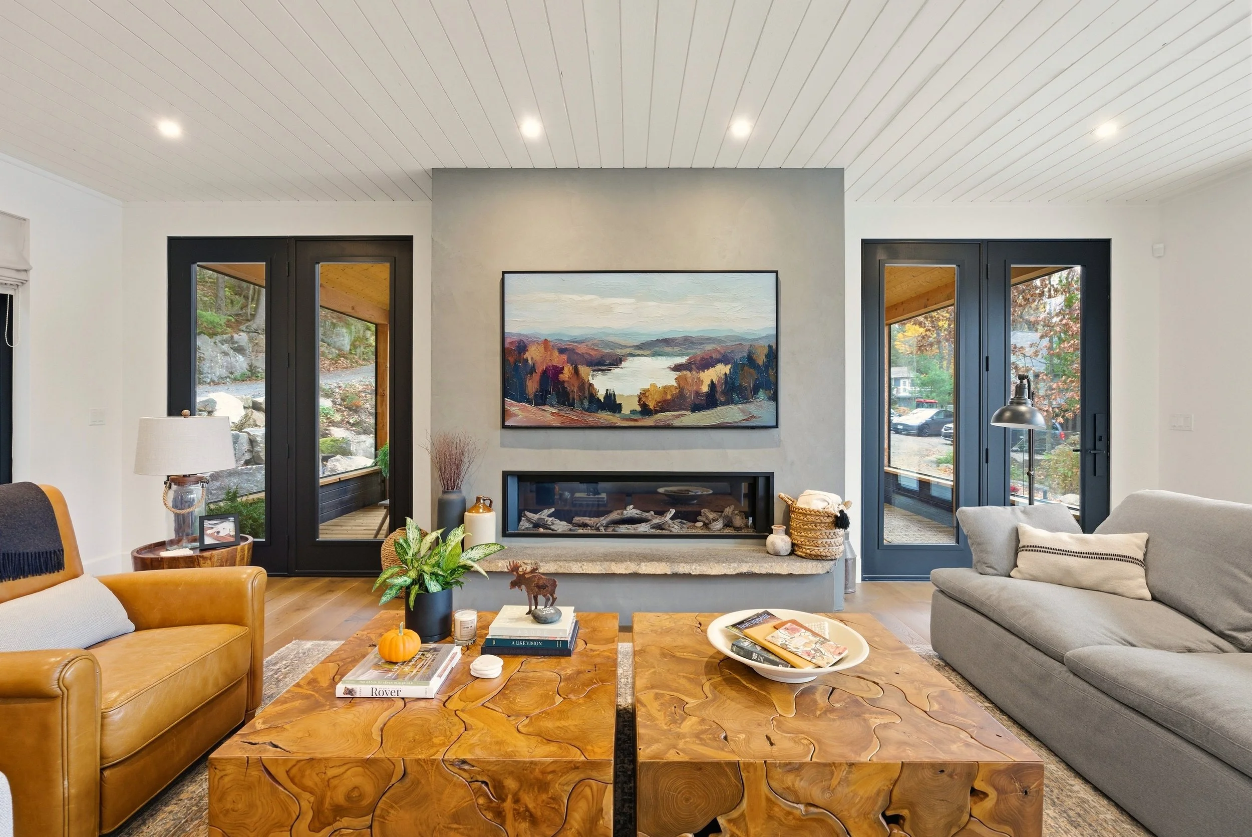 Living room with tan leather sofa, gray sofa, wooden coffee table, fireplace with landscape painting above, large windows, and outdoor view.