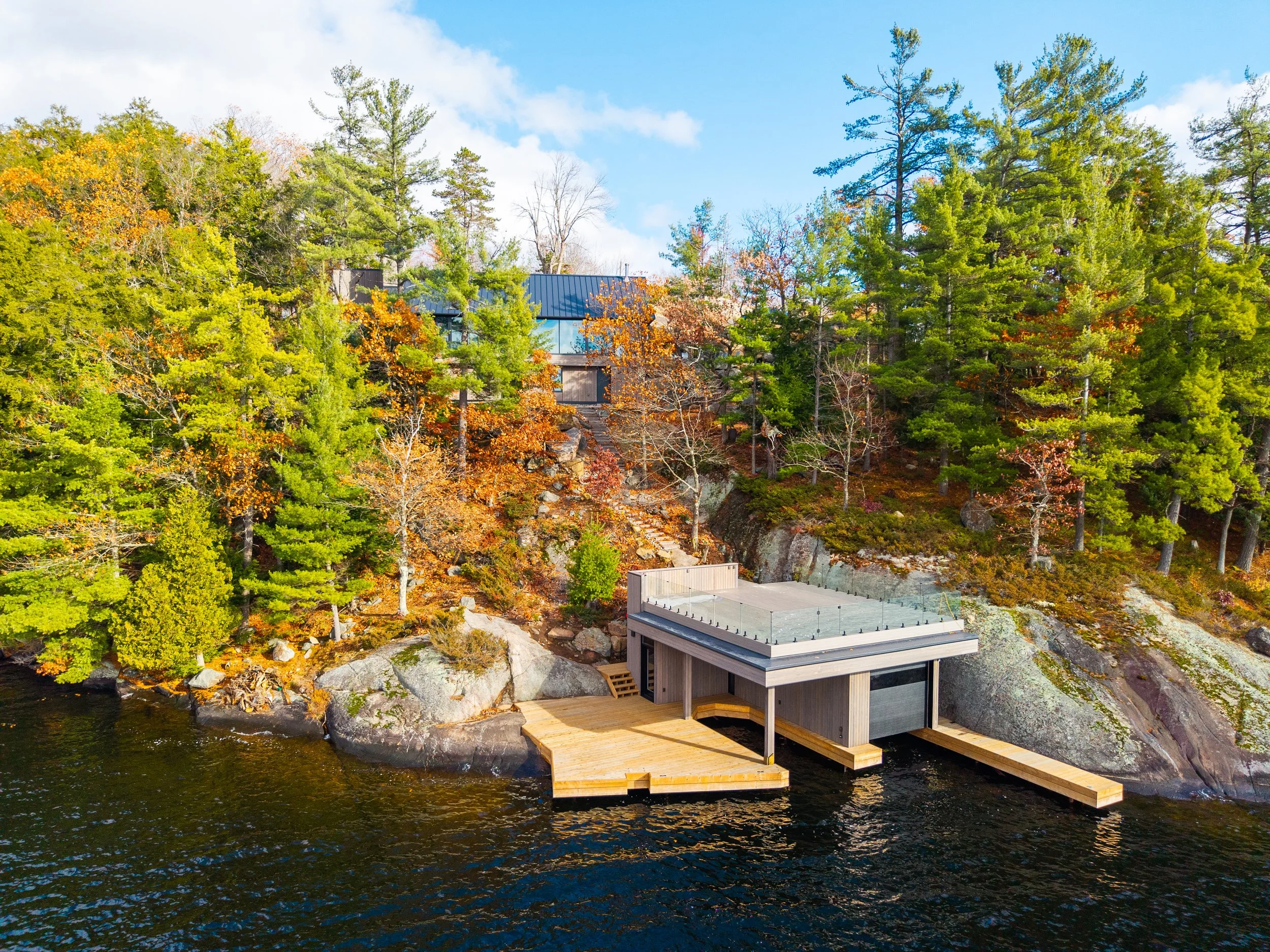 A modern house with a glass balcony situated on a rocky lakeshore, surrounded by tall trees with autumn foliage, under a partly cloudy blue sky.