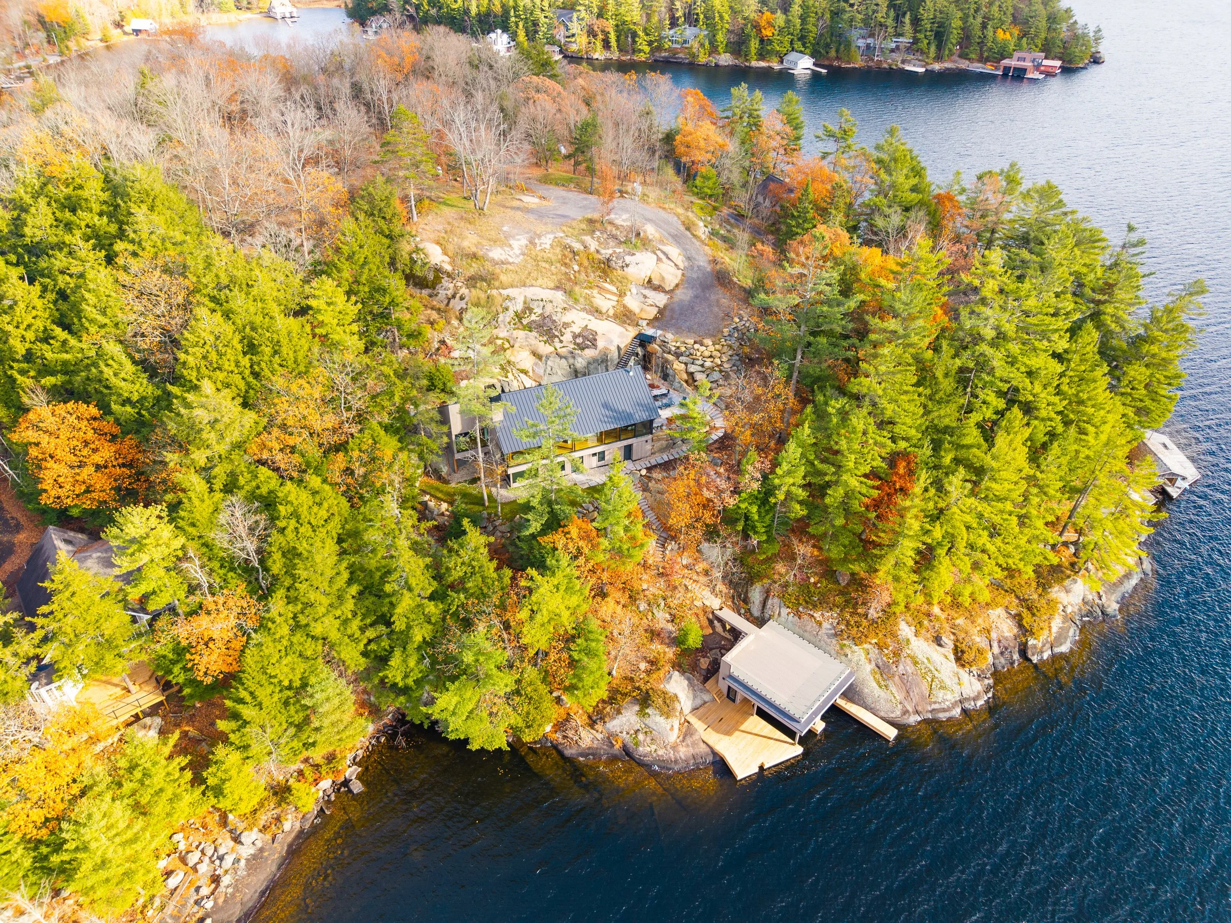 An aerial view of a lakeside property surrounded by autumn trees, with a house, a dock extending into the lake, and additional cabins nearby.