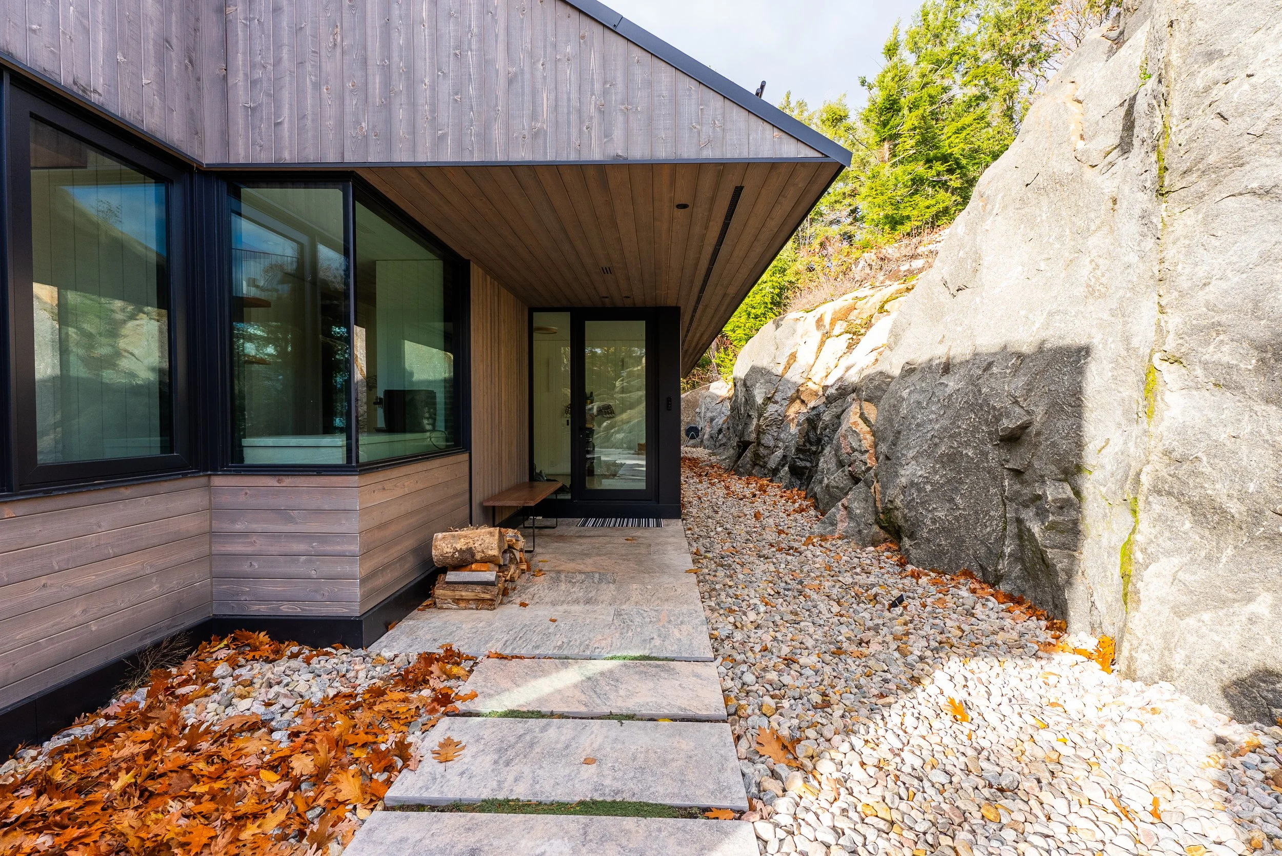 Modern house with wooden exterior, large glass windows, and a small pathway surrounded by rocks and fallen autumn leaves. Large rocky hill on the right side.