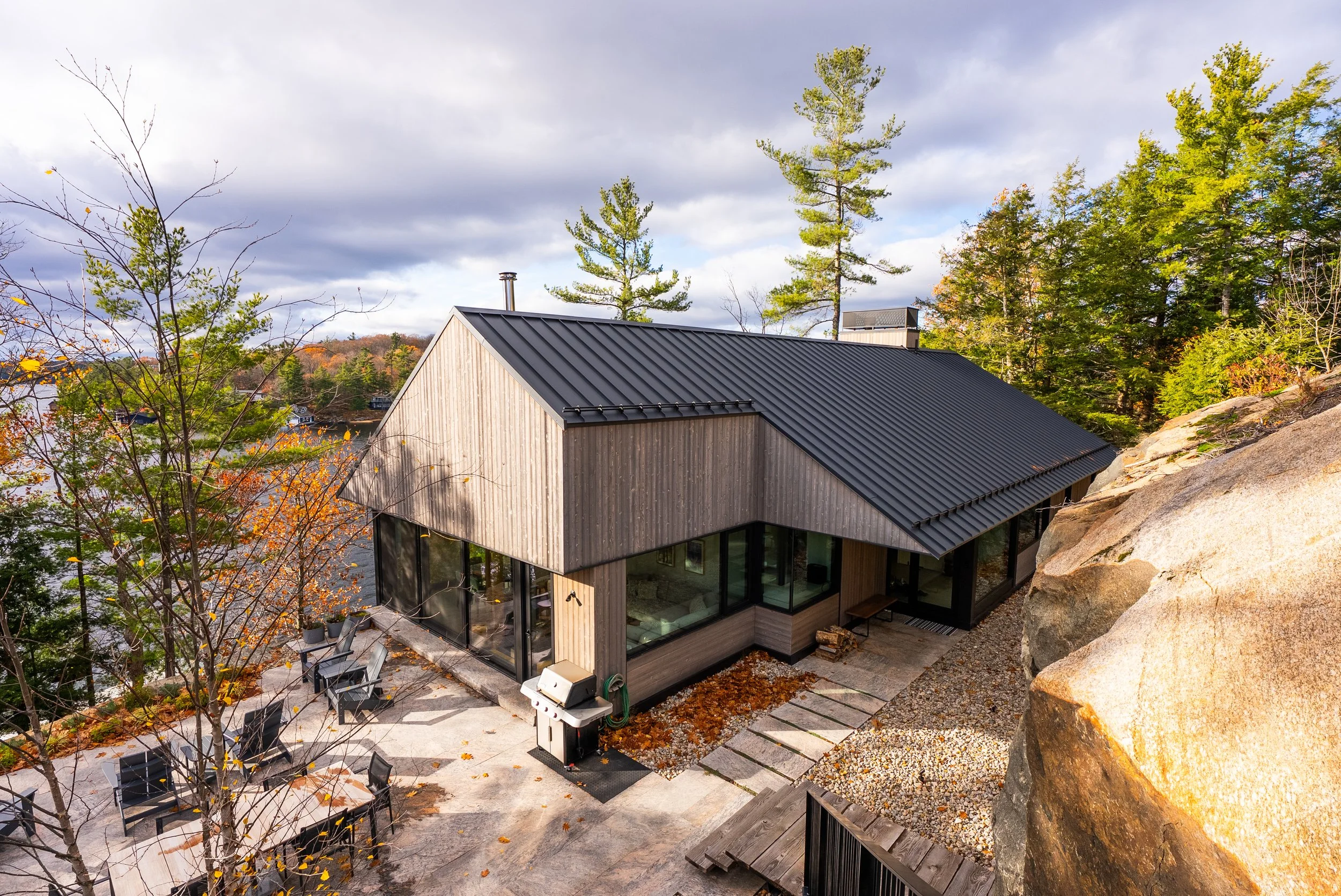 A custom home in Muskoka, built by Conklin's Construction with a black metal roof, large glass windows, and wooden exterior, situated on a rocky hillside surrounded by trees in autumn.