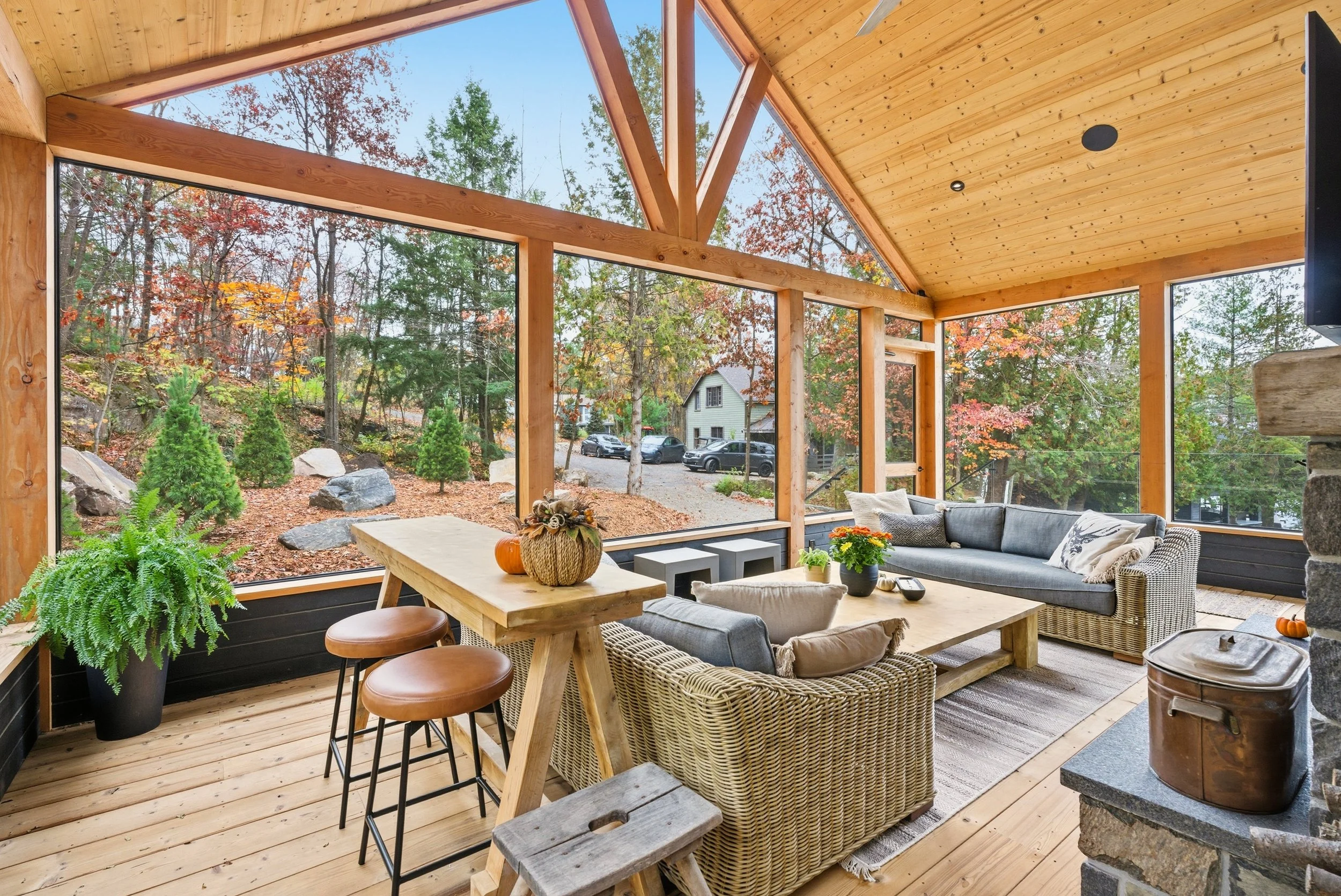 A cozy screened porch with wooden framing, wicker furniture, a coffee table, and large windows showing a wooded yard with autumn leaves.