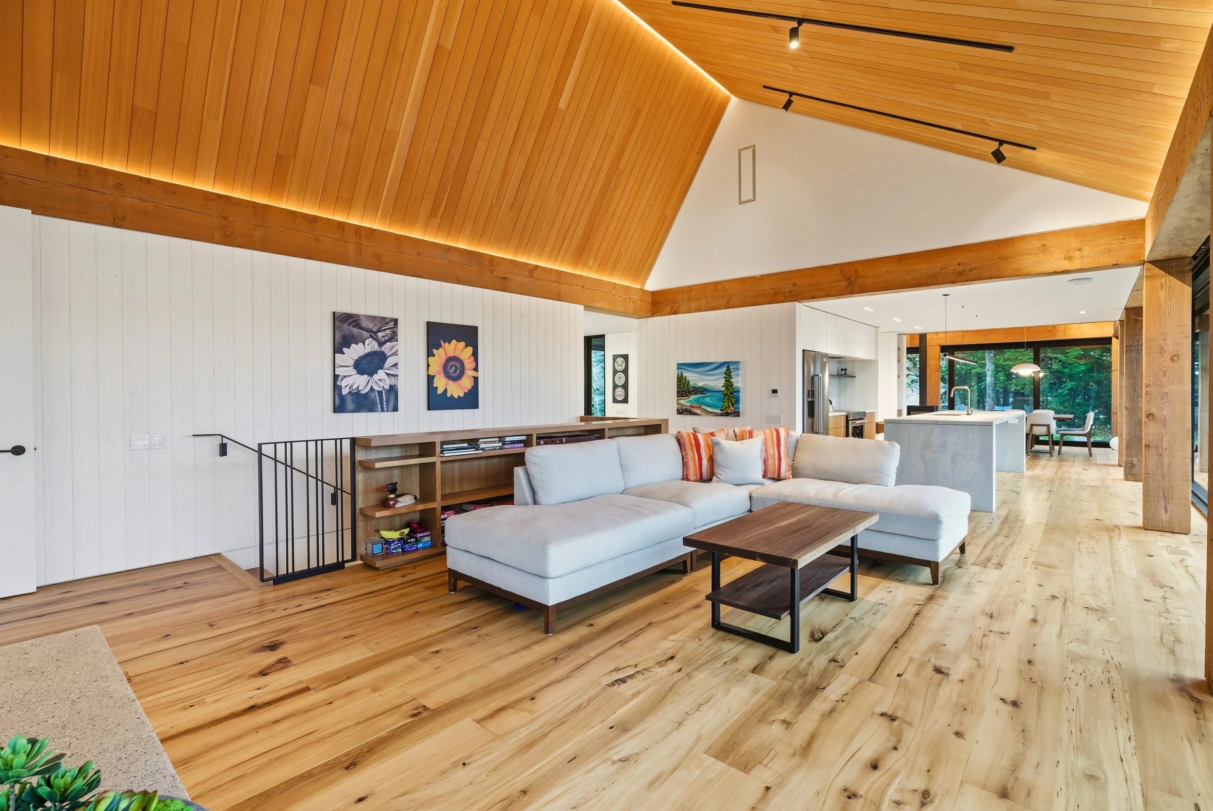Open-concept living room with white sectional sofa, wooden coffee table, and wall art in a house with vaulted wooden ceiling and large windows showing greenery outside.