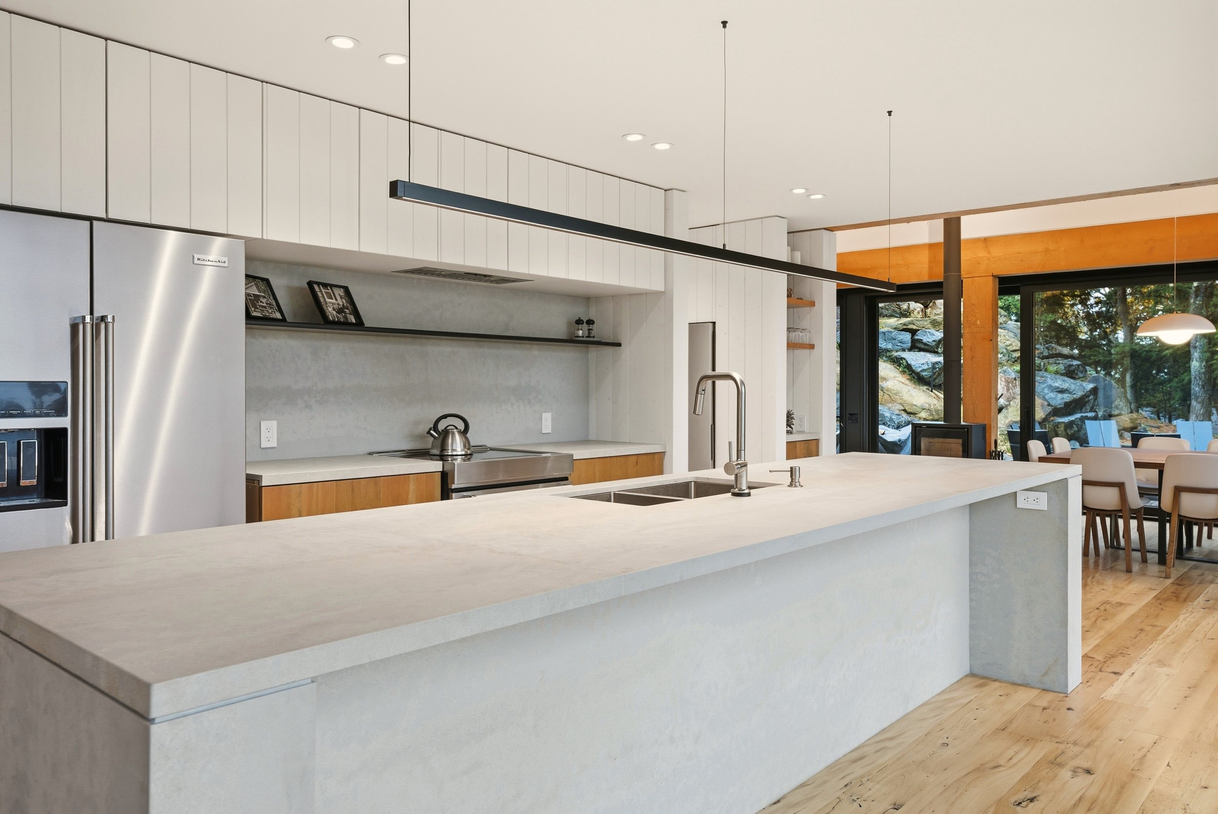 Modern kitchen with white marble island, stainless steel appliances, and wood accents, with a dining area and large windows showing an outdoor landscape.