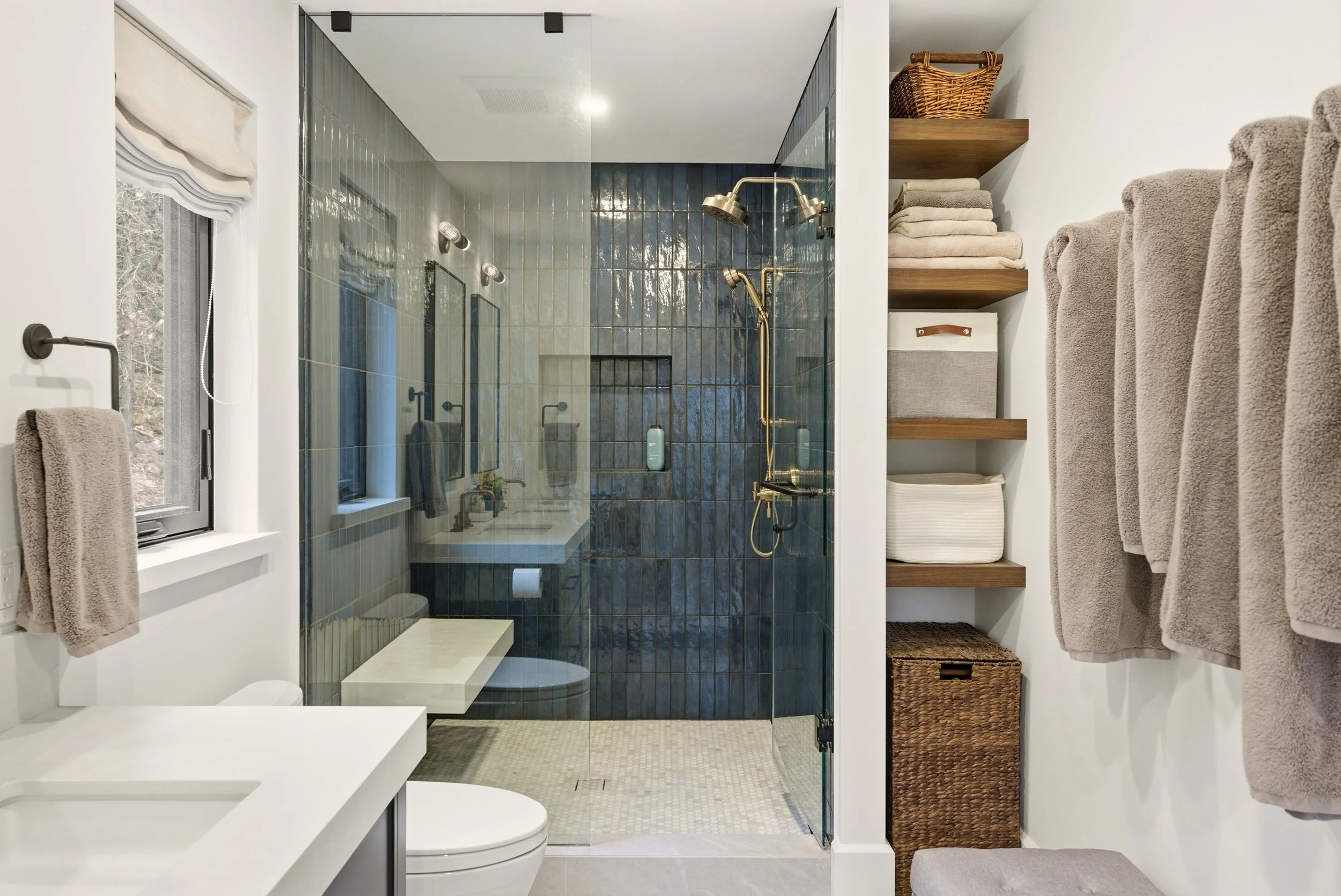 Bathroom with walk-in shower enclosed by glass, with dark blue tiles, gold fixtures, and a window with beige roman shade. White vanity with a beige towel on a towel rack, open shelving with folded towels, baskets, and storage boxes, and neutral-color