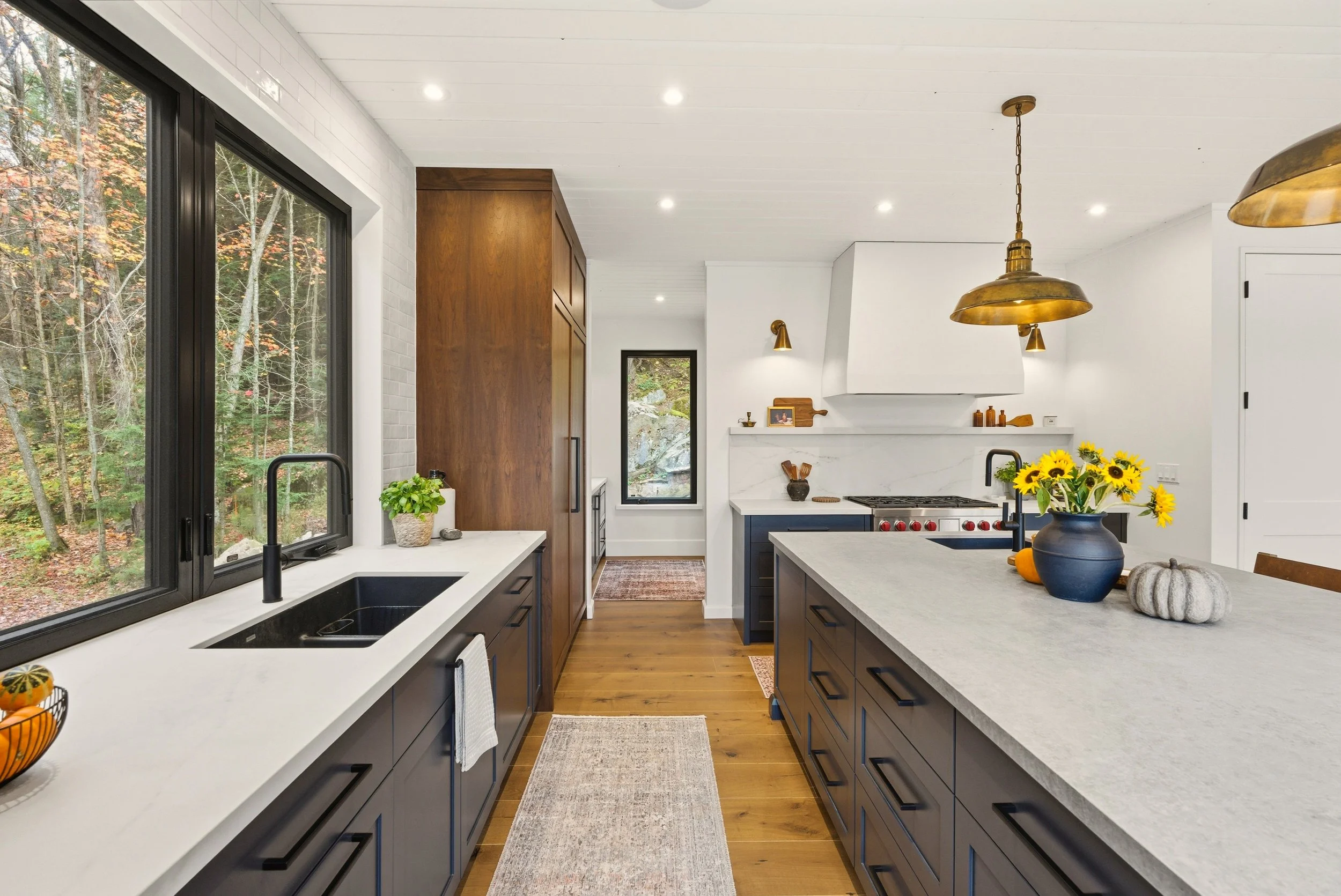 Modern kitchen with white walls, wooden accents, and large windows overlooking a wooded area. Features navy blue cabinets, a large white countertop island with a vase of sunflowers, gold pendant lights, and farmhouse-style sink.