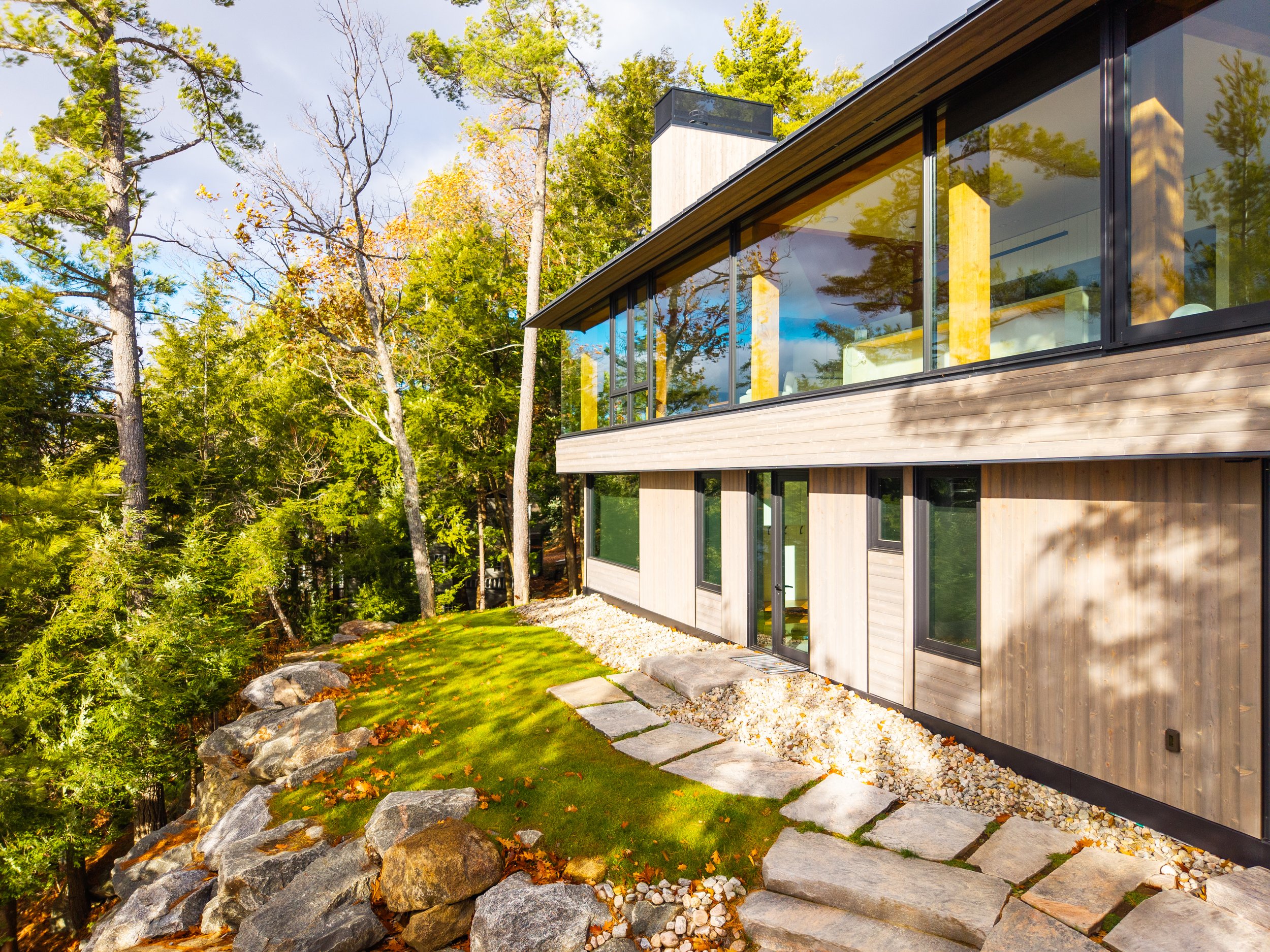 Modern house with large glass windows and wooden exterior, surrounded by green trees and a landscaped yard with rocks and stepping stones.
