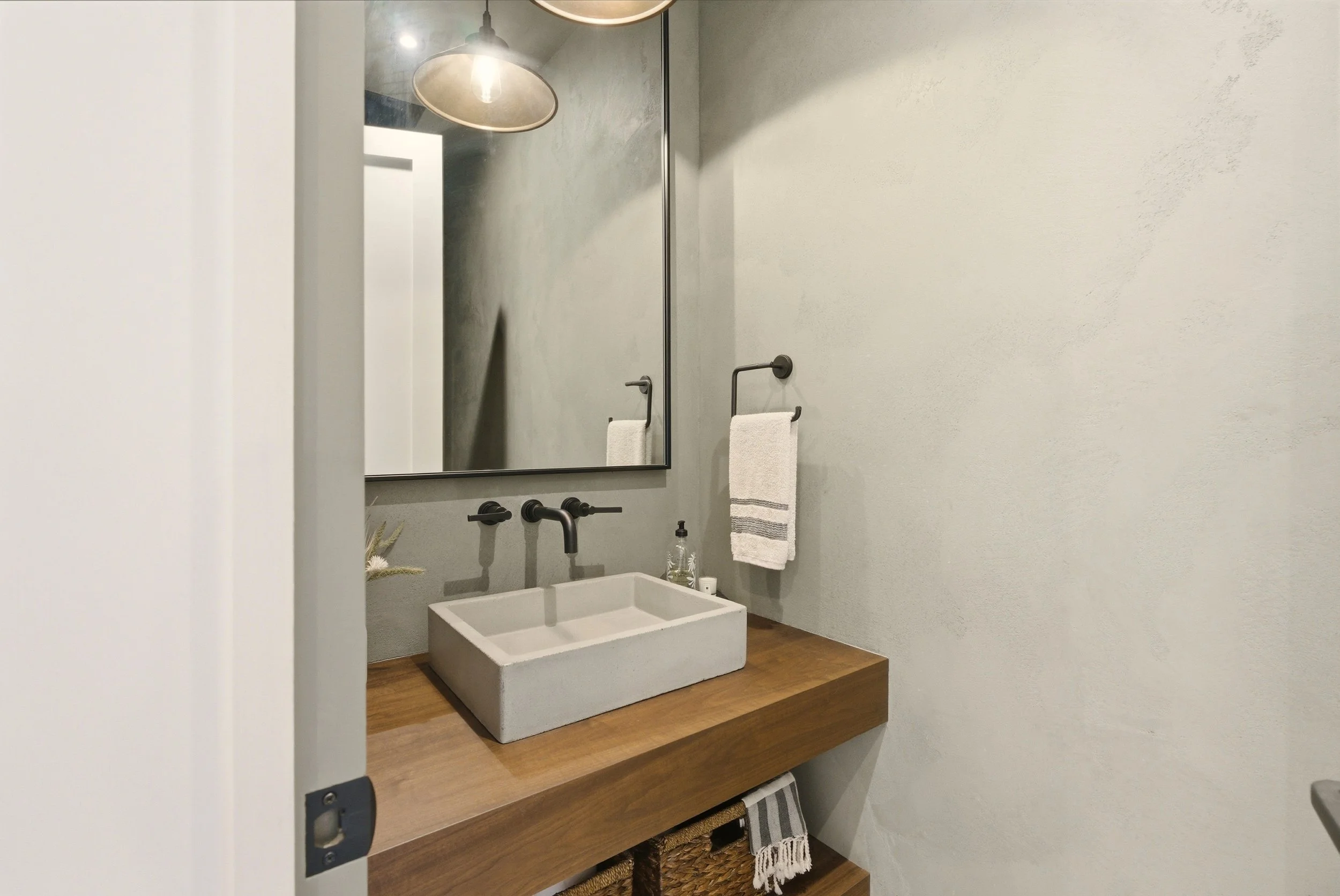 Modern bathroom sink with a concrete basin, wooden countertop, large rectangular mirror, and black fixtures, with towels hanging on black wall-mounted towel racks.