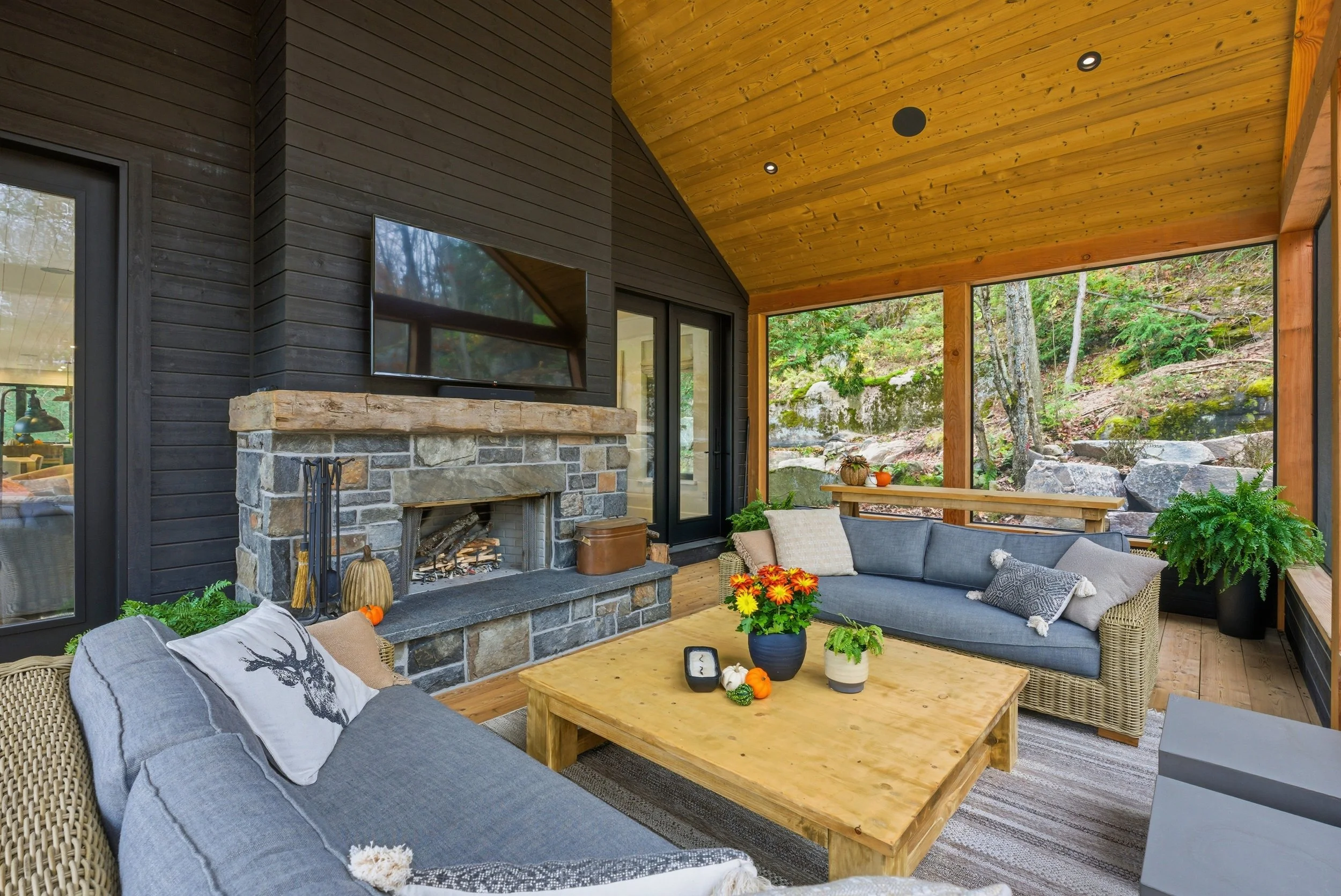 Cozy screened-in porch with wooden ceiling and walls, featuring a stone fireplace with a mounted TV, wicker and fabric sofas with decorative pillows, a wooden coffee table decorated with potted plants and pumpkins, and a view of a wooded outdoor land