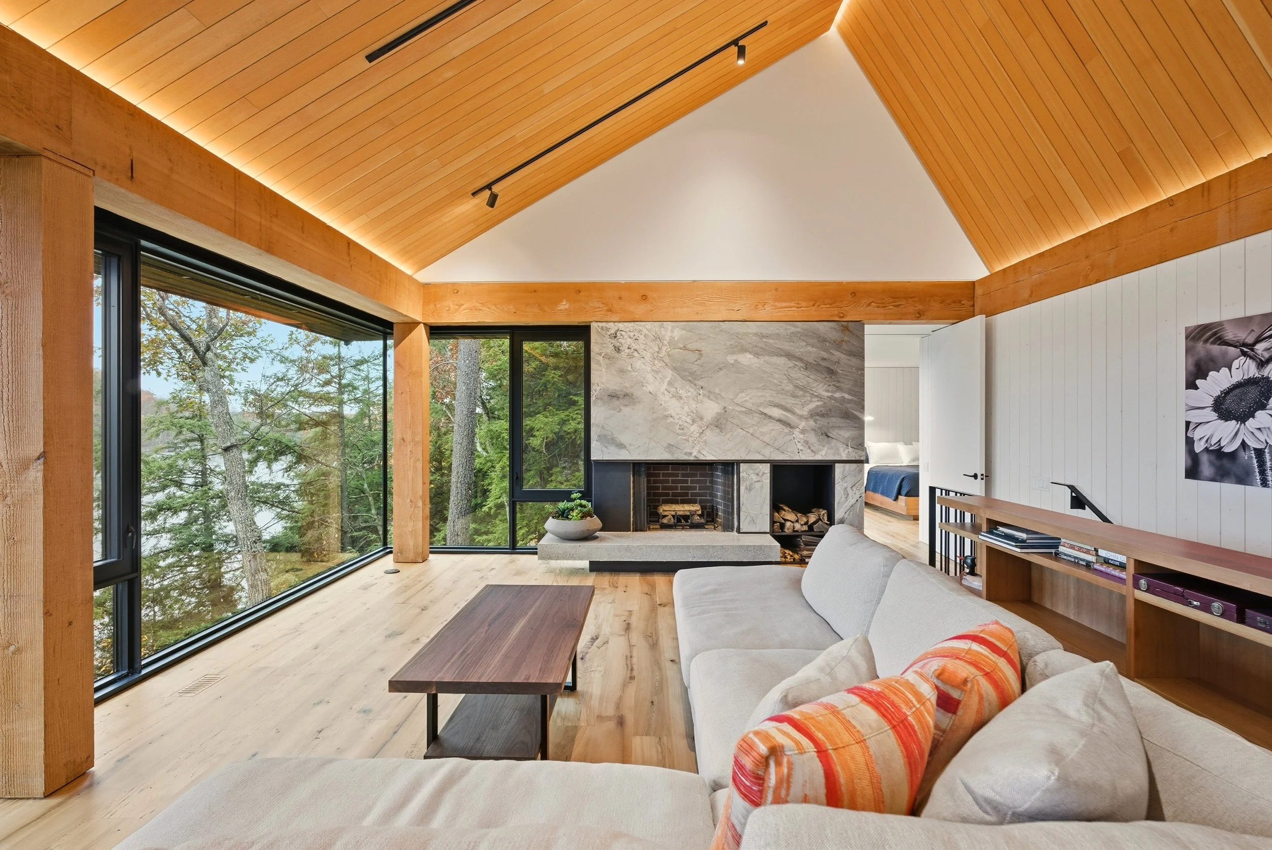 Living room with large windows showing trees outside, a modern fireplace with gray marble surround, beige sofa with orange and beige cushions, wooden ceiling and floors, and a view into a bedroom.