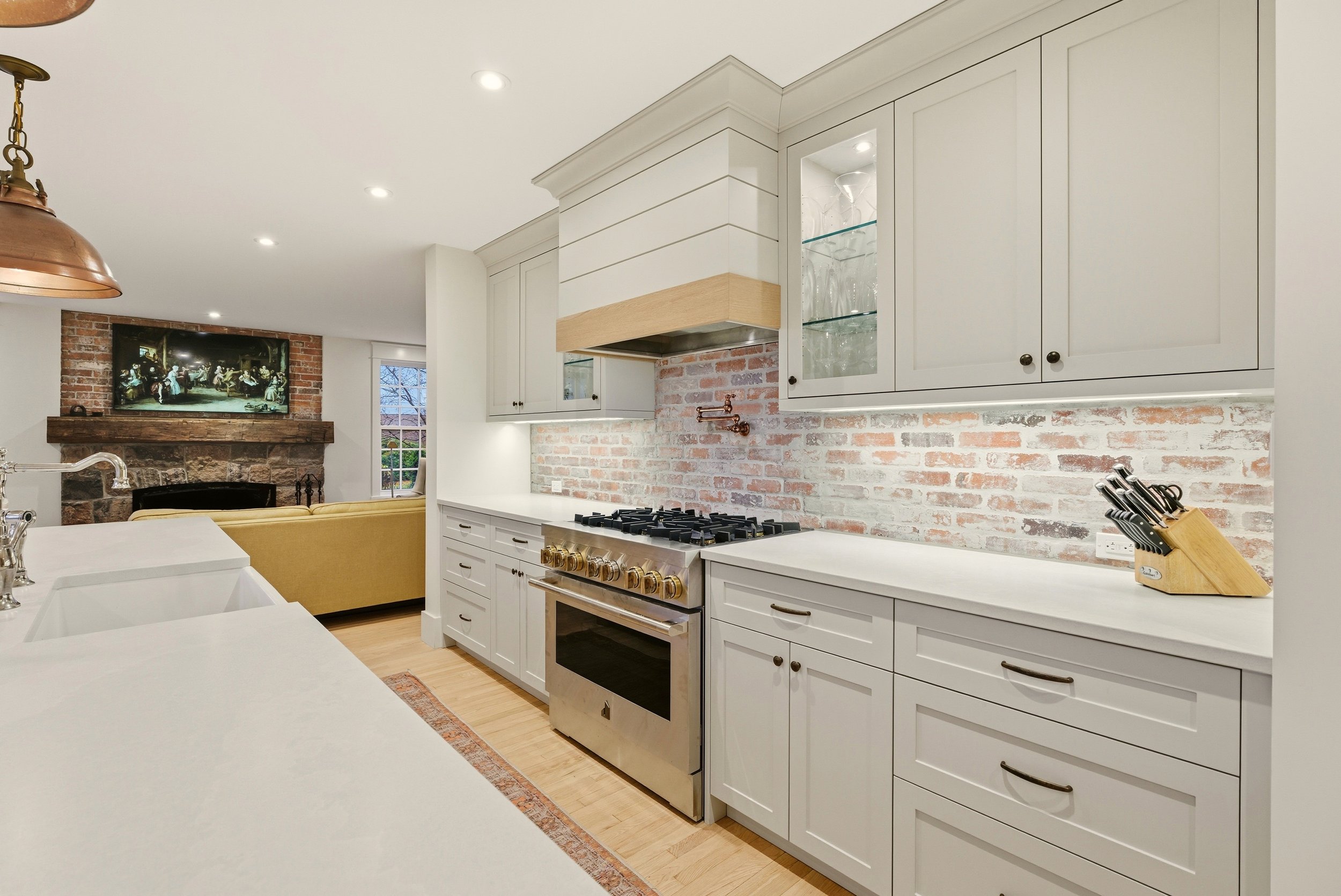 Modern kitchen with white cabinets, brick backsplash, stainless steel stove, wooden countertops, and a view into a cozy living room with a brick fireplace and a flat-screen TV.