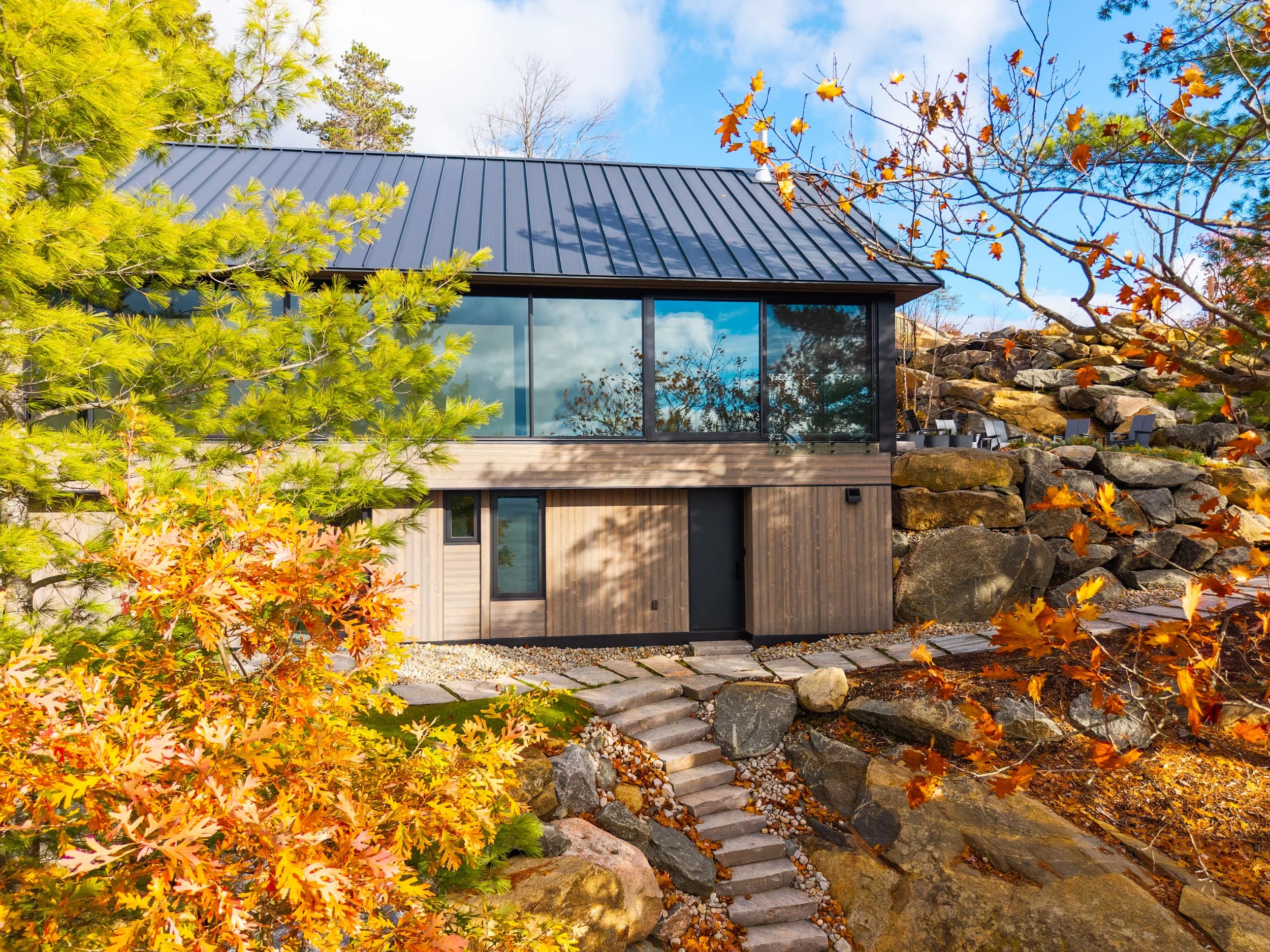 Modern house with large glass windows and a metal roof surrounded by autumn trees and rocks.