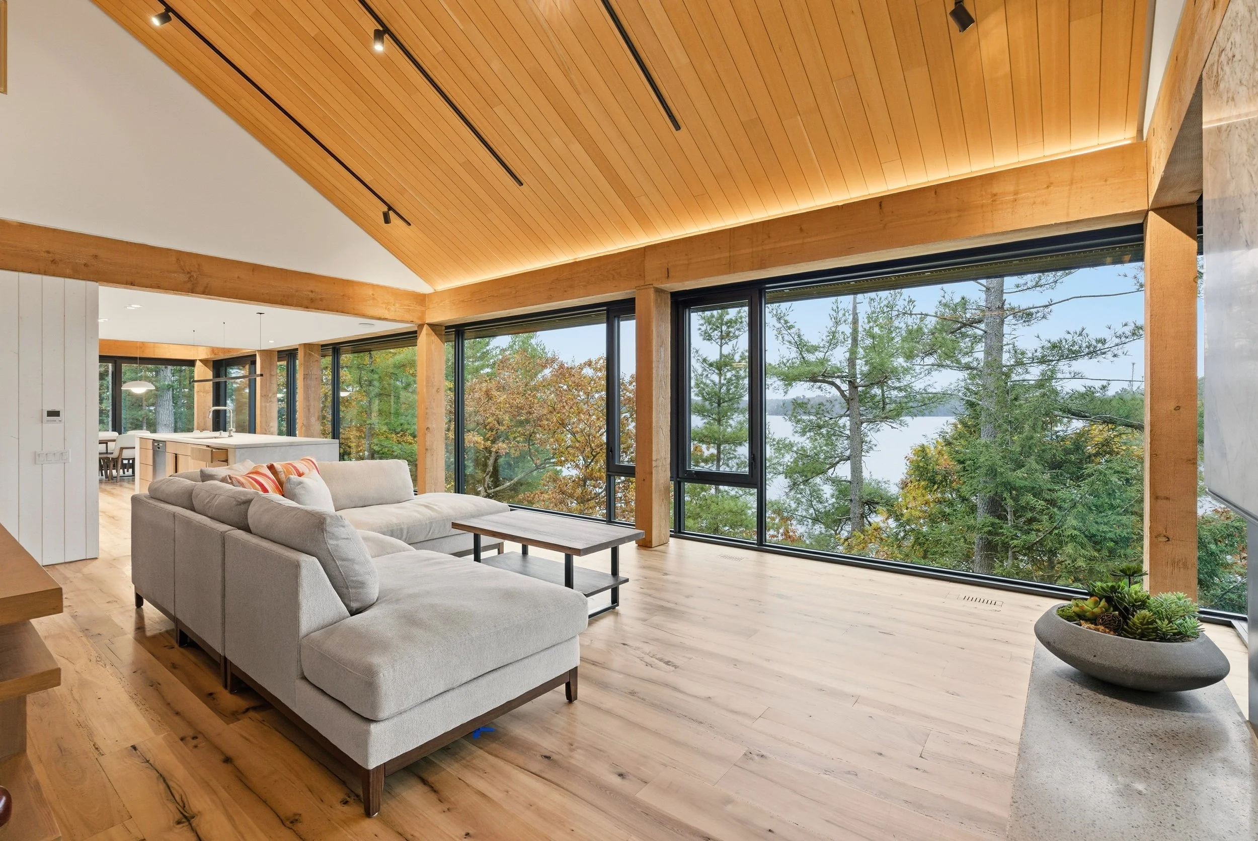 Modern living room with large windows overlooking trees, a white sectional sofa, a wooden coffee table, and wooden ceiling beams.