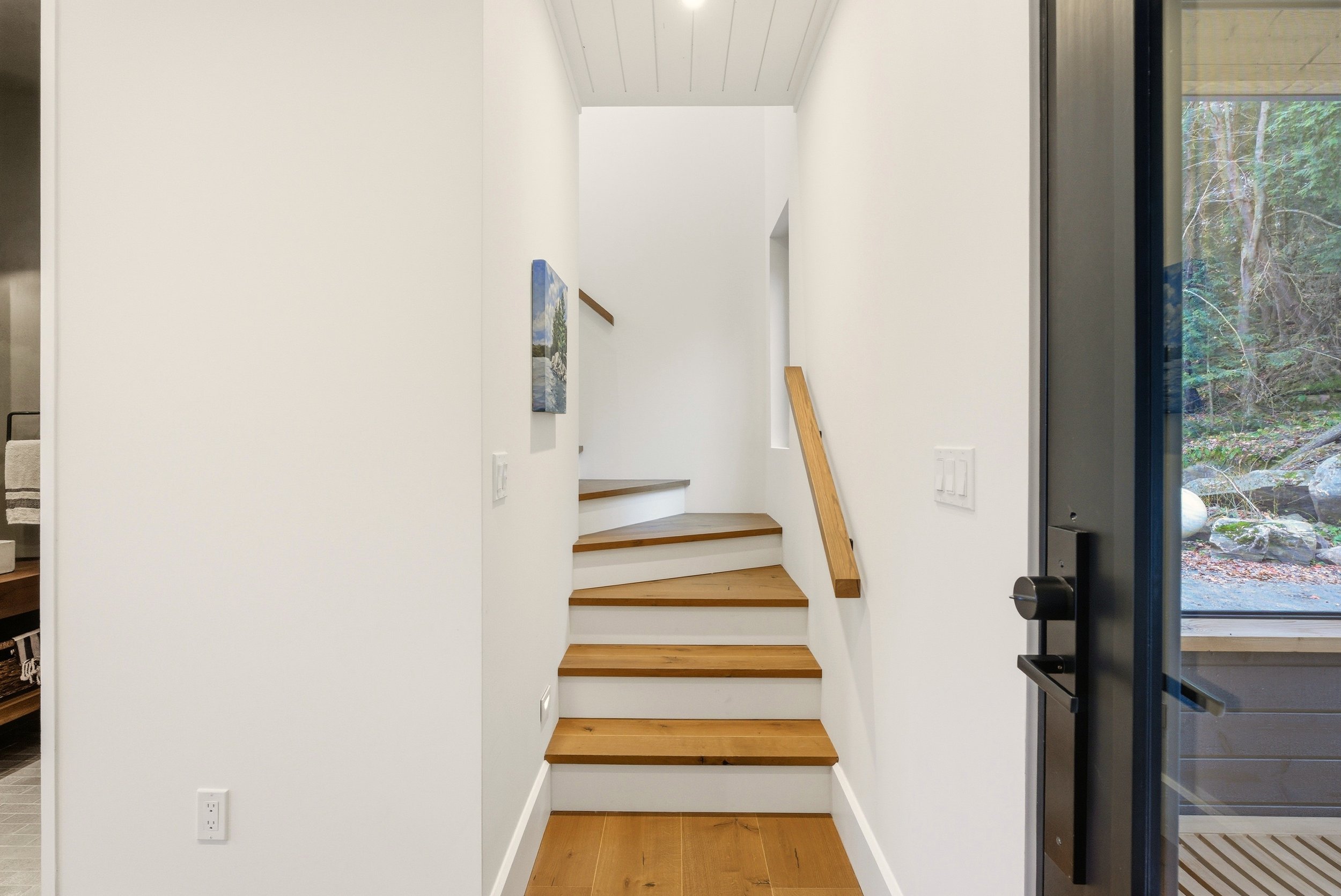 Interior view of a staircase with wooden steps and white walls, next to a glass door showing a wooded outdoor scene.
