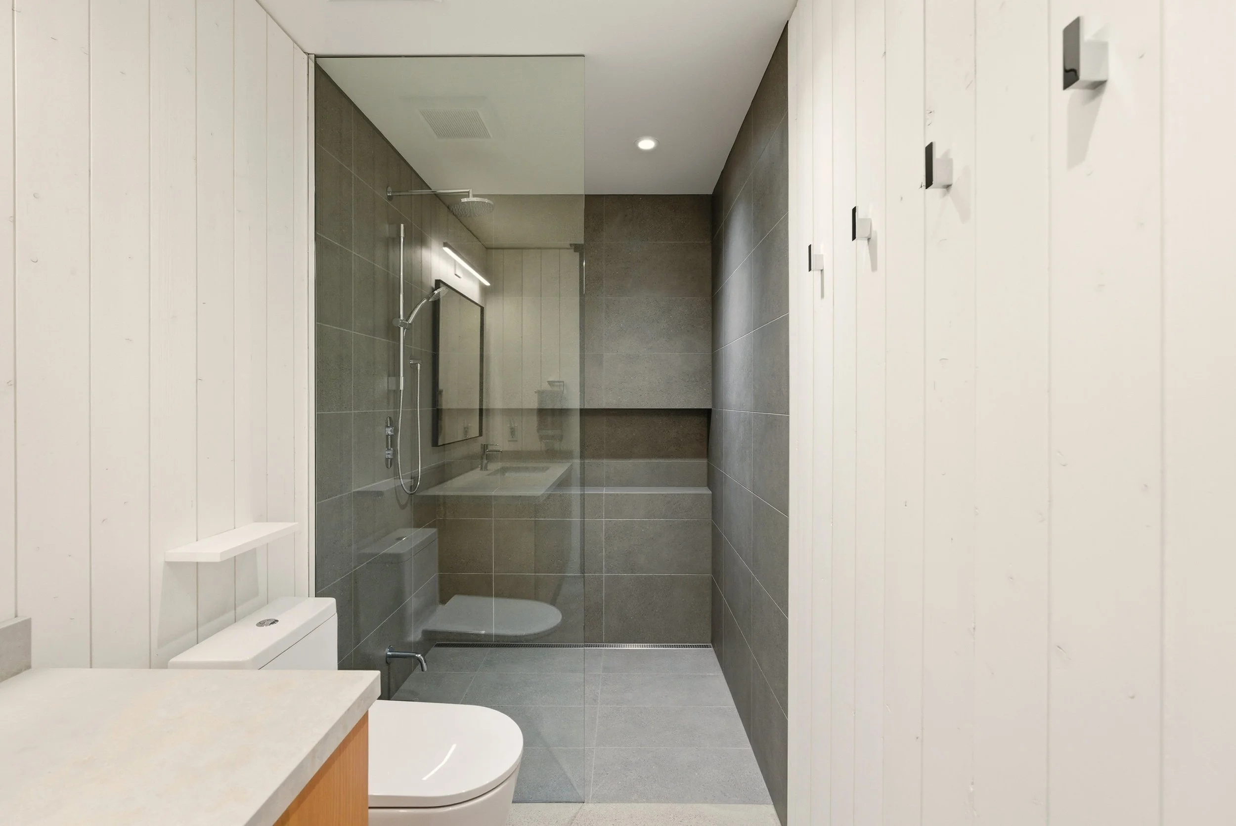 Modern bathroom with white paneled walls, gray tiled shower area with glass partition, wall-mounted shower head, mirror, toilet, and wooden countertop.