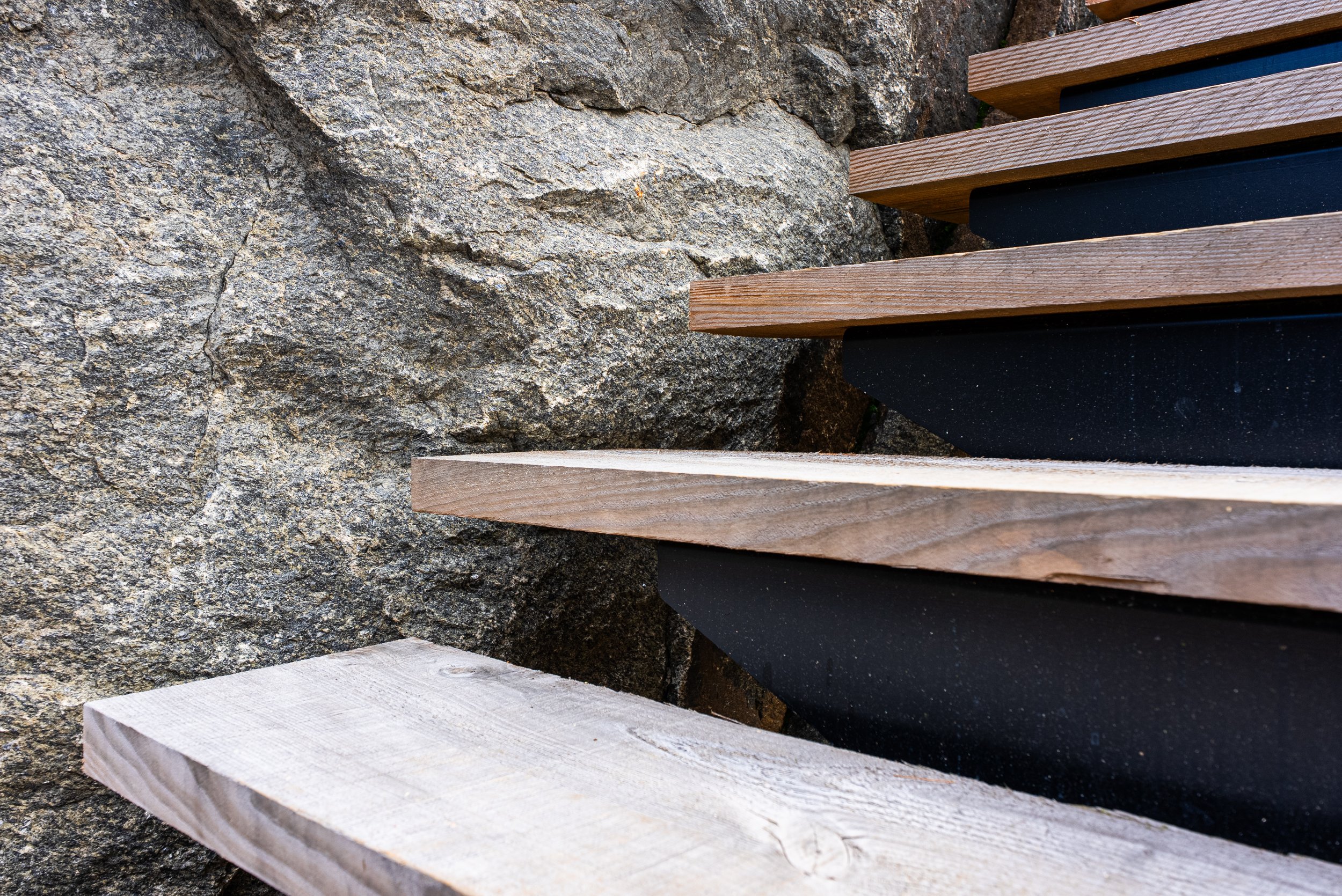 Close-up of wooden stairs with black supports next to a textured rock wall.
