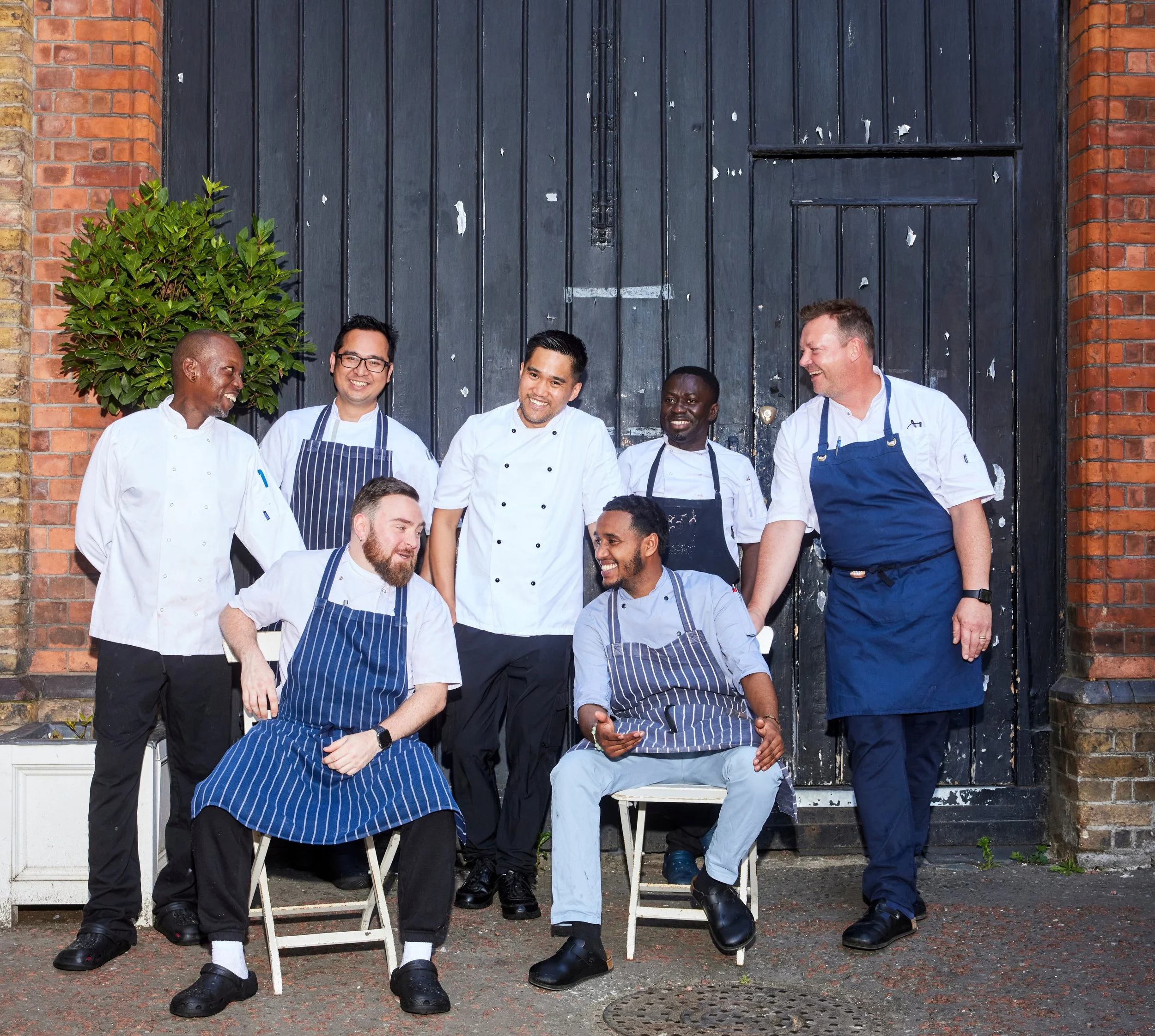 Group of eight chefs and restaurant staff laughing and talking outside a building with a black wooden door, brick wall, and a potted plant.