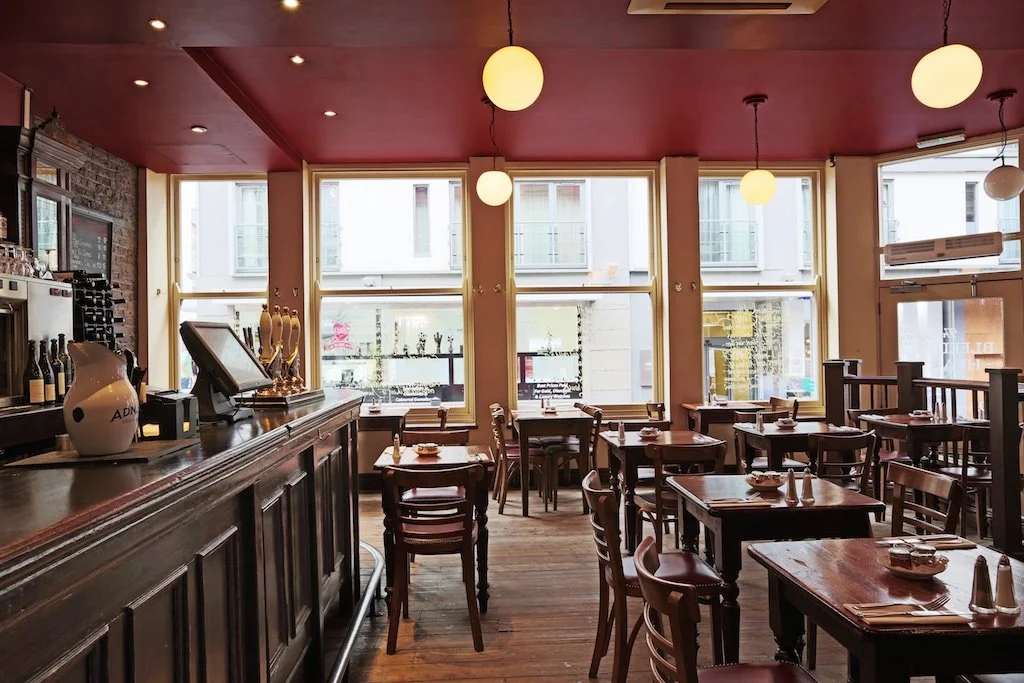 Interior of a cozy restaurant with wooden tables and chairs, large front windows, a bar counter on the left with bottles and a pitcher, and hanging round lights on the ceiling.