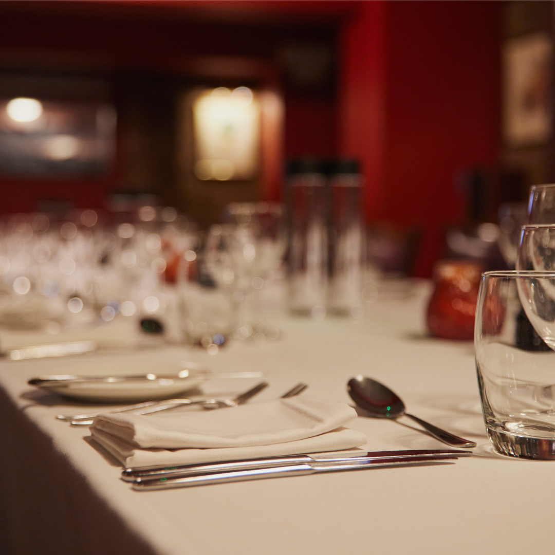 Close-up of a dinner table setting with silverware, a folded napkin, a glass of water, and blurred wine glasses in the background in a restaurant.