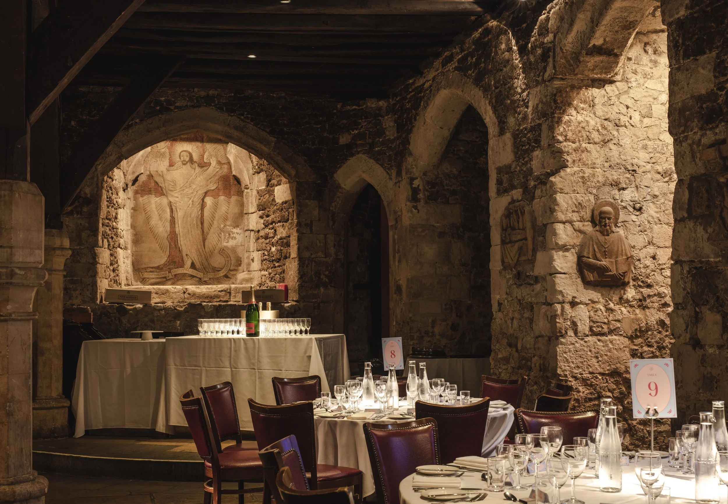 Elegant restaurant with stone walls, arched doorways, and religious artwork. Round tables covered with white tablecloths, set with glassware and silverware, and wooden chairs. One table has a sign with the number 9, and the bar area has bottles and g
