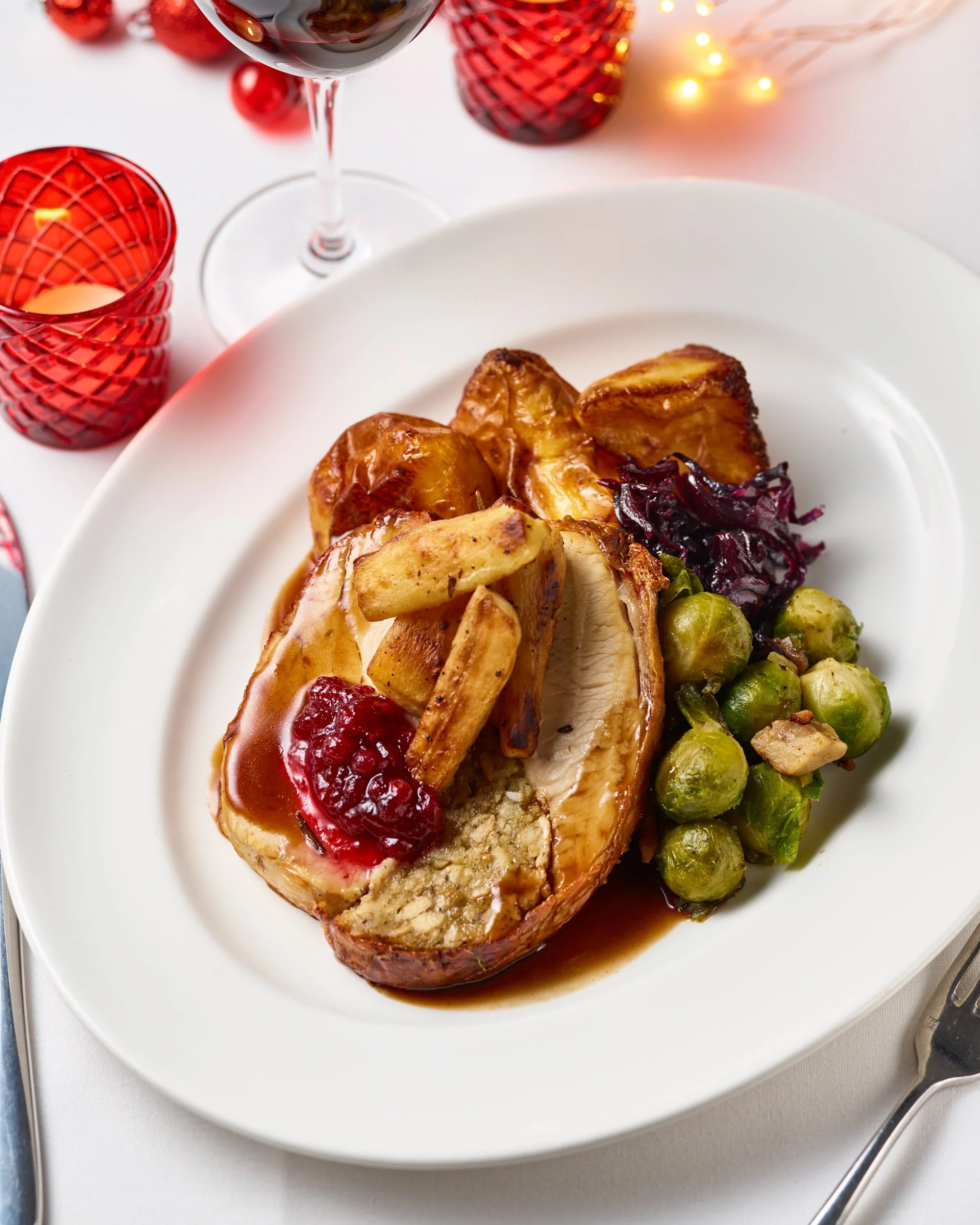 Holiday ham with cooked potatoes, Brussels sprouts, and red cabbage, served on a white plate with wine and holiday decor in the background.