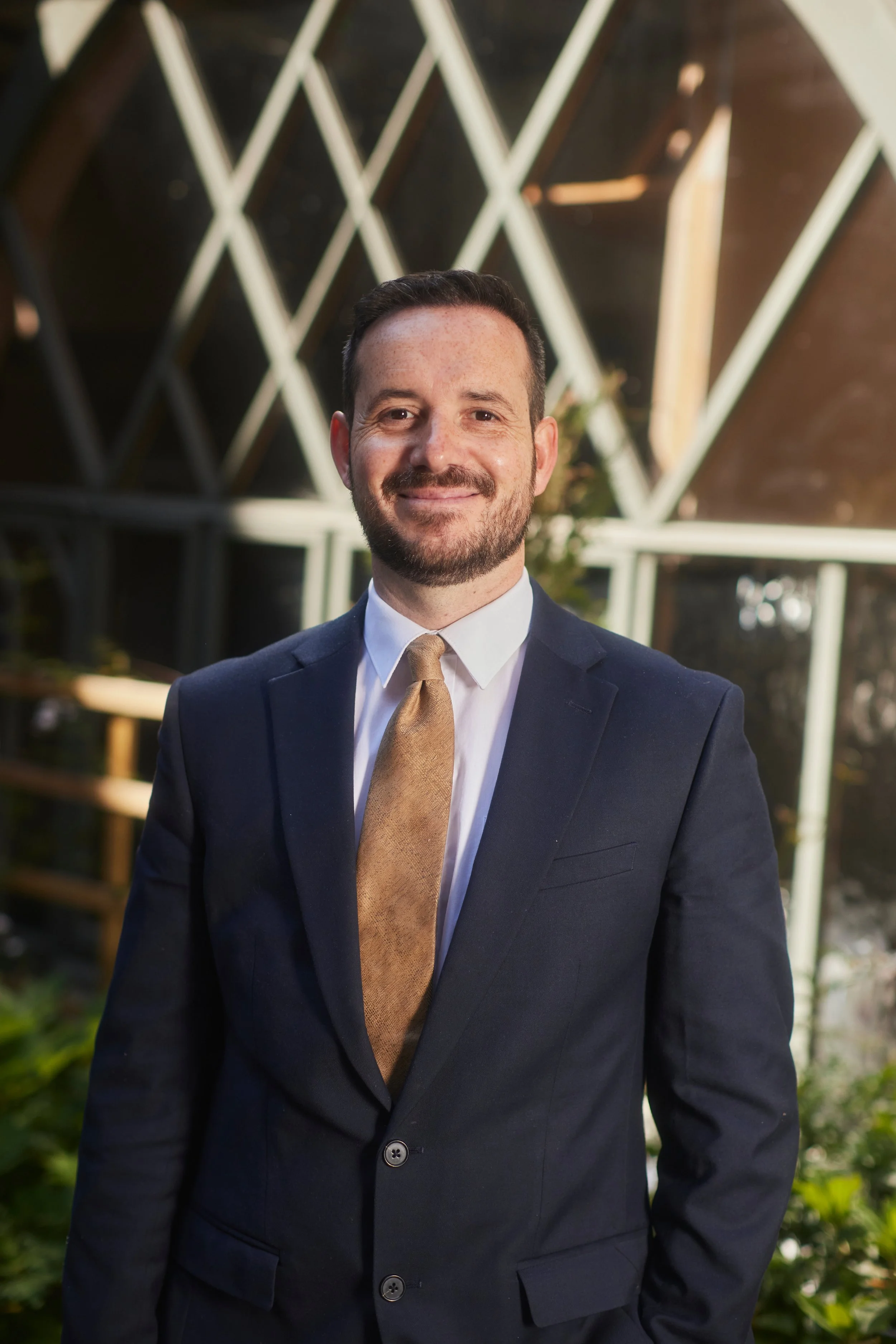 A man in a dark suit and light brown tie standing outside with a geometric metallic structure in the background.