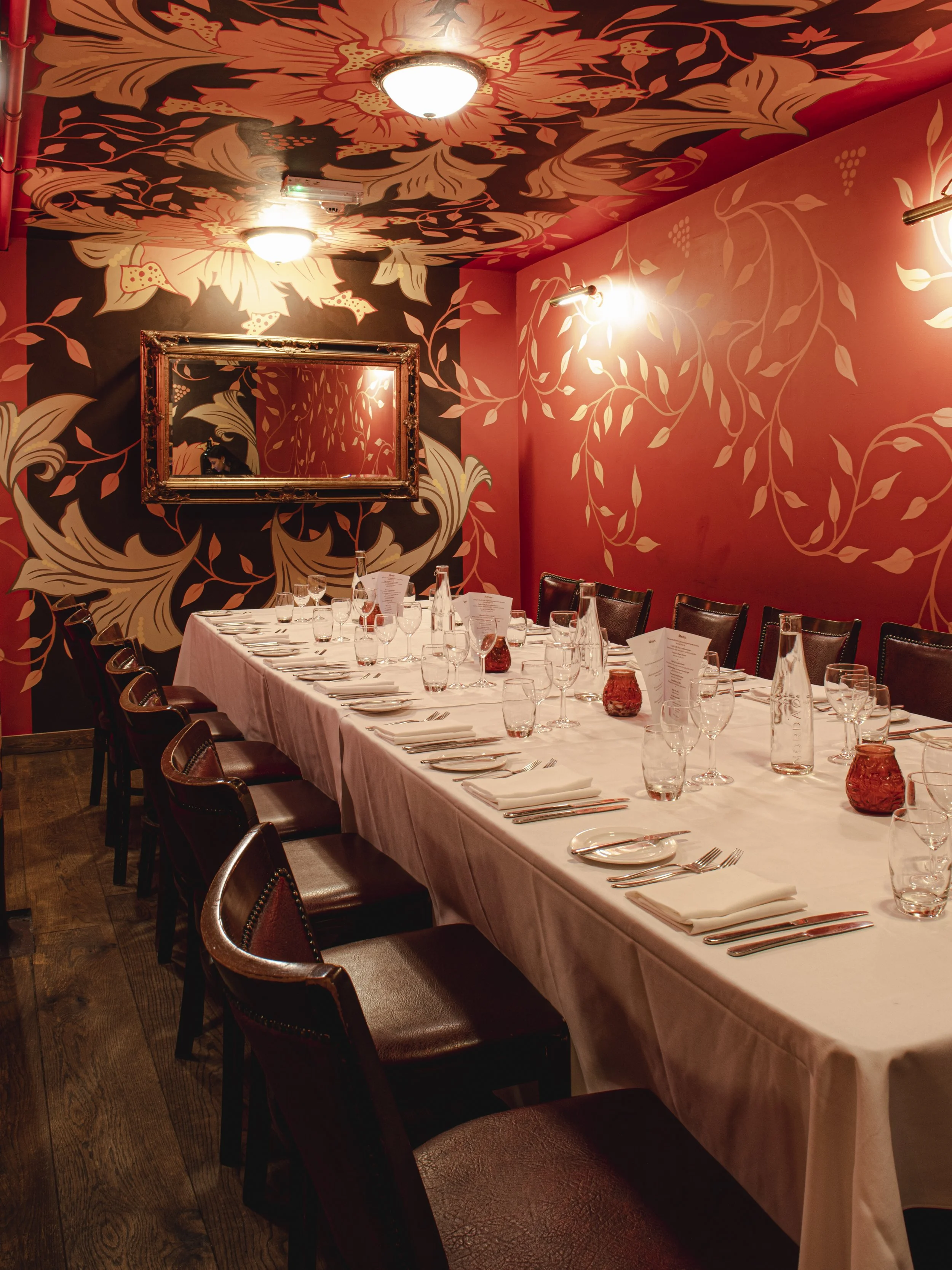 A long rectangular dining table set for a formal dinner in an elegant room with pink and black floral wallpaper and framed mirror on the wall. The table has white tablecloth, glassware, plates, silverware, napkins, and small red candle holders.