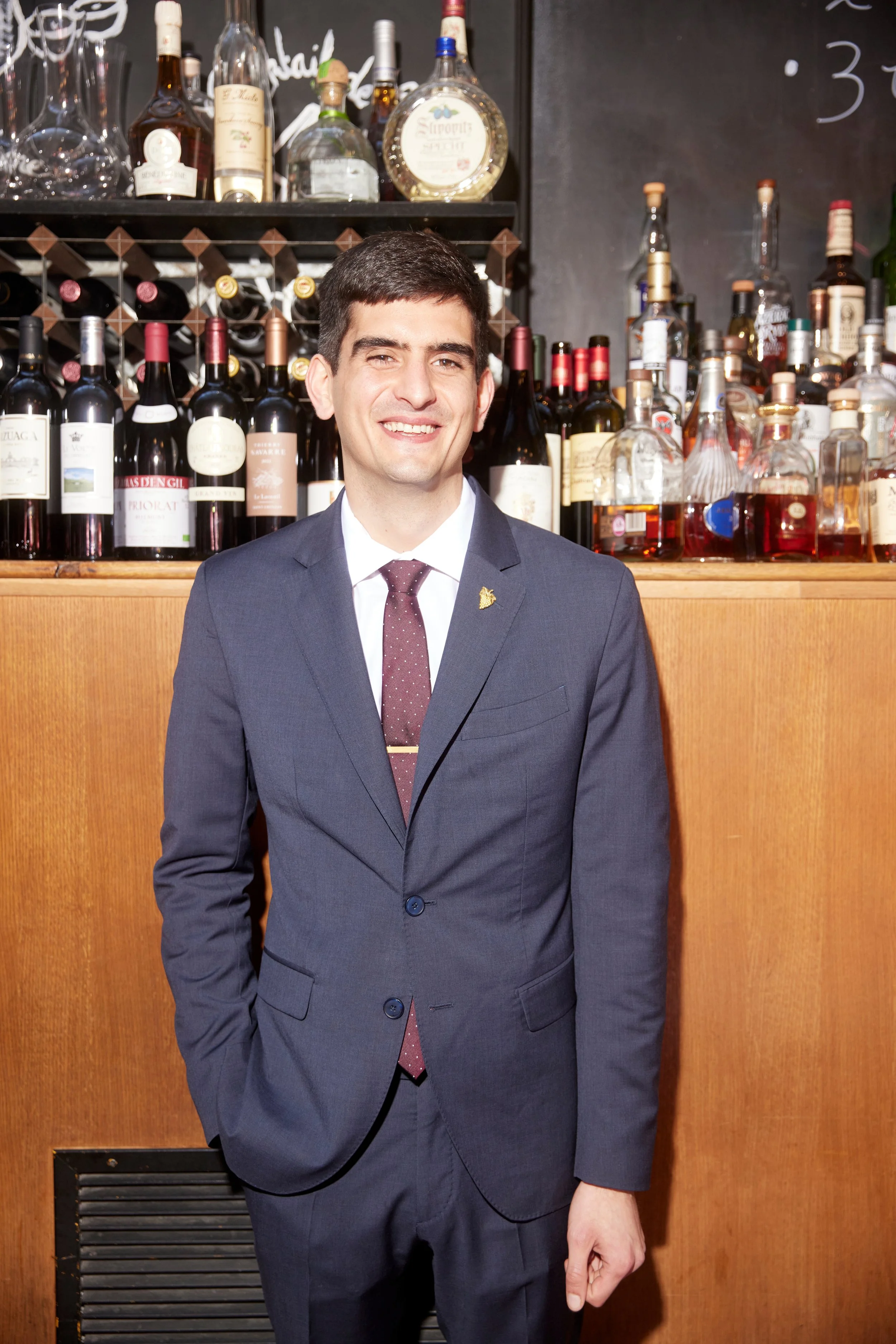 Man dressed in a dark suit, white shirt, and patterned tie, standing in front of a backlit bar with various bottles of wine and liquor bottles on the shelves behind him, smiling.