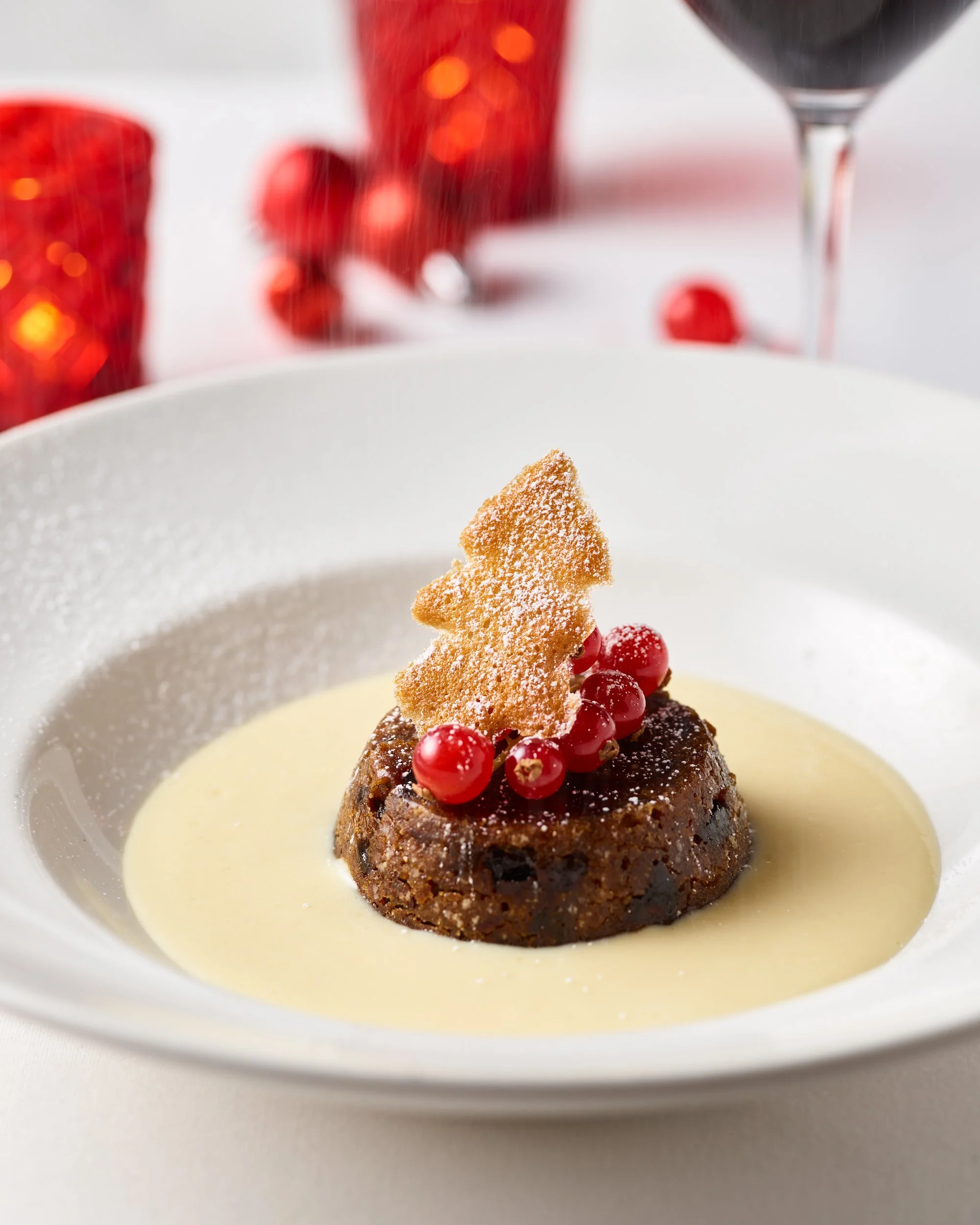 Festive dessert with a round cake topped with red berries and a crispy shard, served in a white bowl with a creamy sauce, with red holiday decorations and wine in the background.