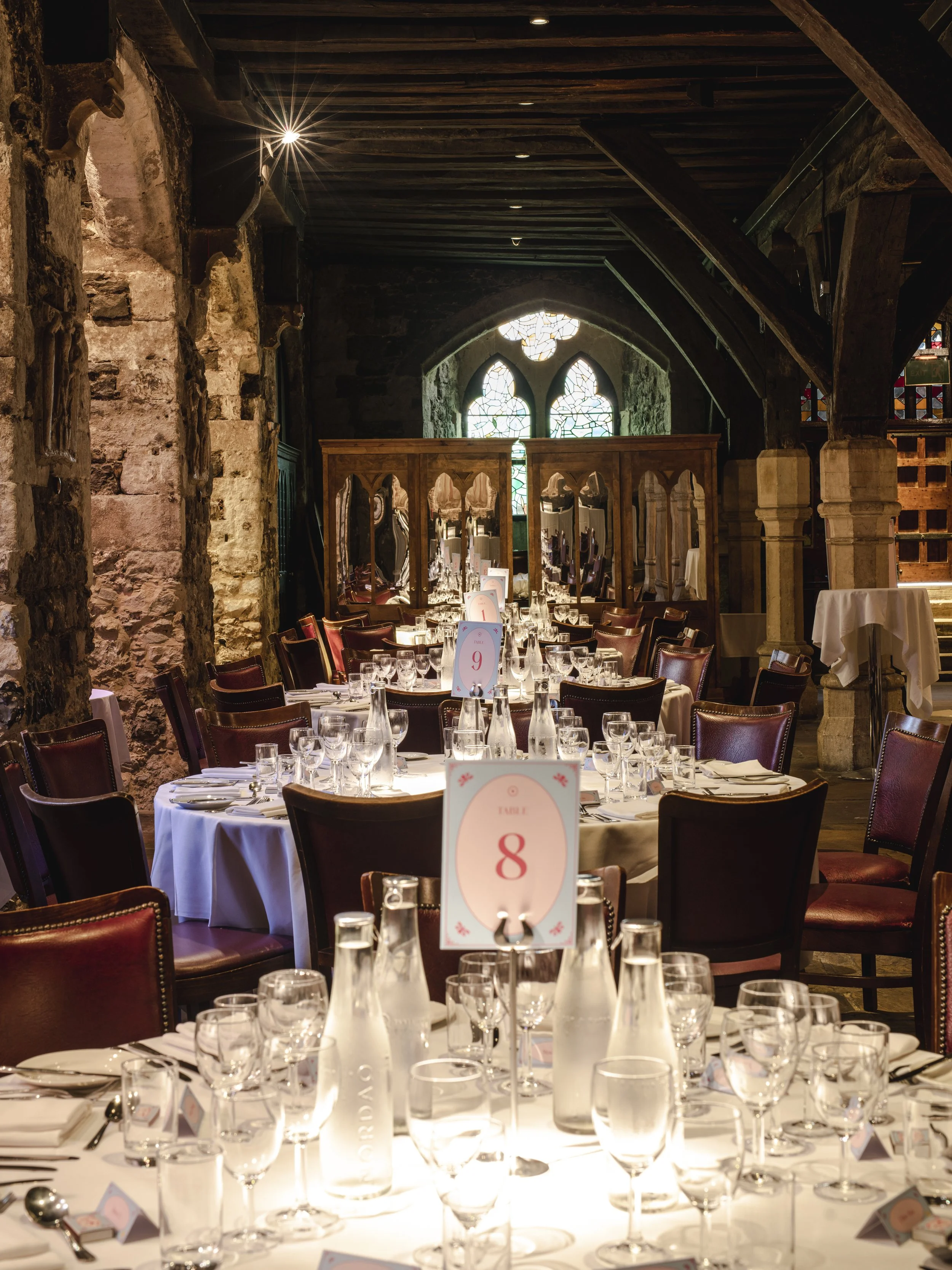 Elegant dining setup with tables covered in white tablecloths, surrounded by wooden chairs with maroon upholstery in a rustic stone and wood interior, featuring stained glass windows at the back.