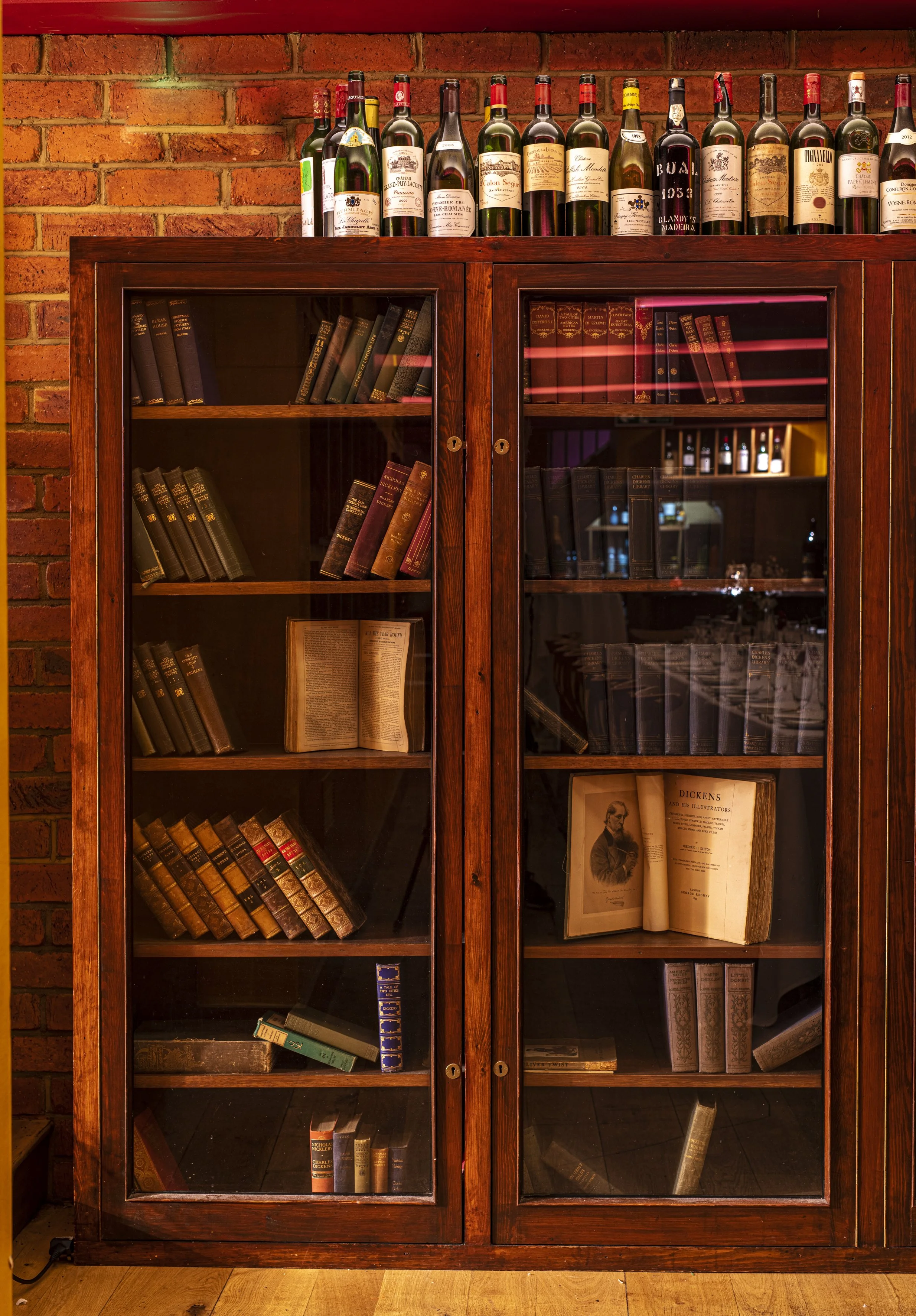 A wooden bookshelf with glass doors containing old books. Several bottles of wine are arranged on top of the bookshelf against a brick wall.