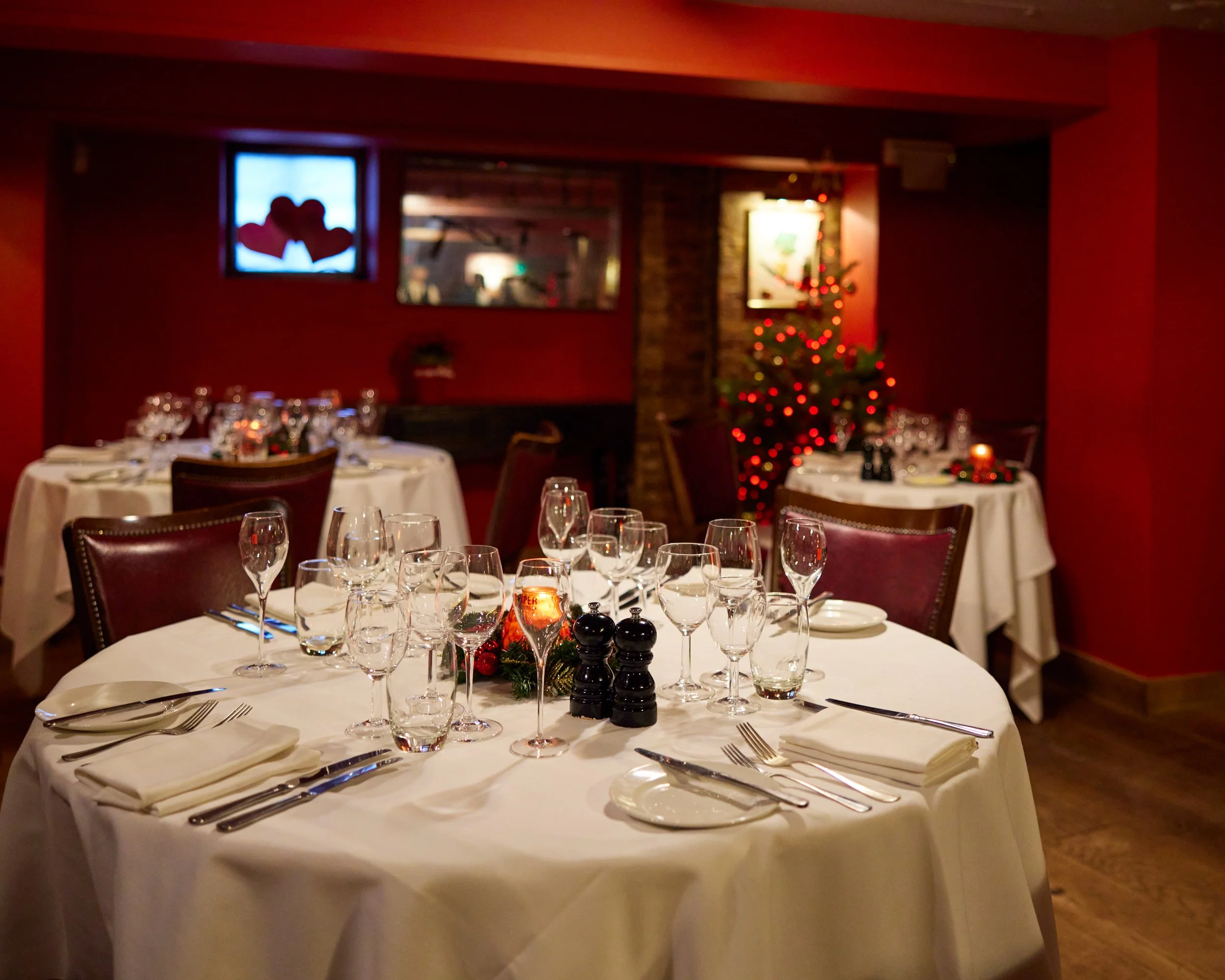 A cozy restaurant dining area decorated for Christmas with round tables set with white tablecloths, glassware, silverware, and napkins. There is a small Christmas tree with red lights and ornaments in the background, along with festive decorations an