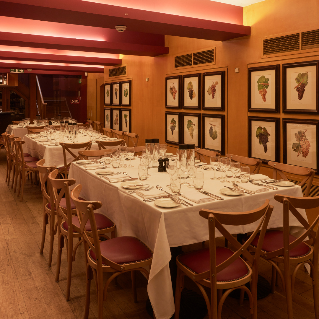 A dining room with long tables covered in white tablecloths, set with glassware, plates, and silverware, surrounded by wooden chairs with red cushions, and framed grapevine artwork on the wood-paneled walls, under pinkish ceiling lights.