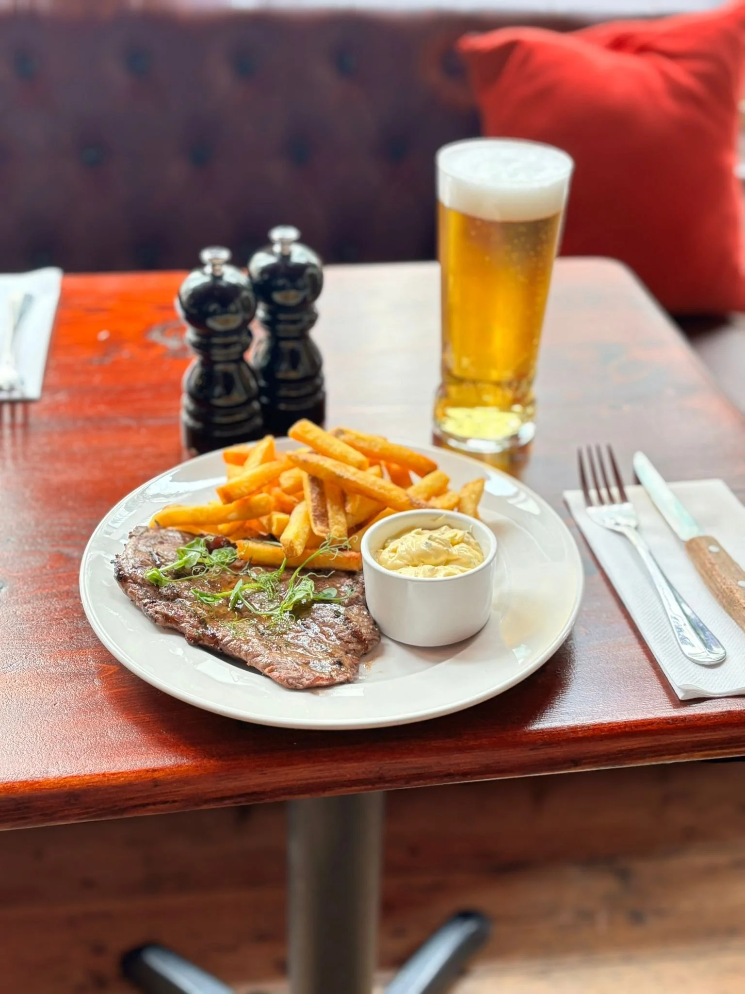 Plate with steak, French fries, and a cup of creamy sauce, with a glass of beer in the background on a wooden table.