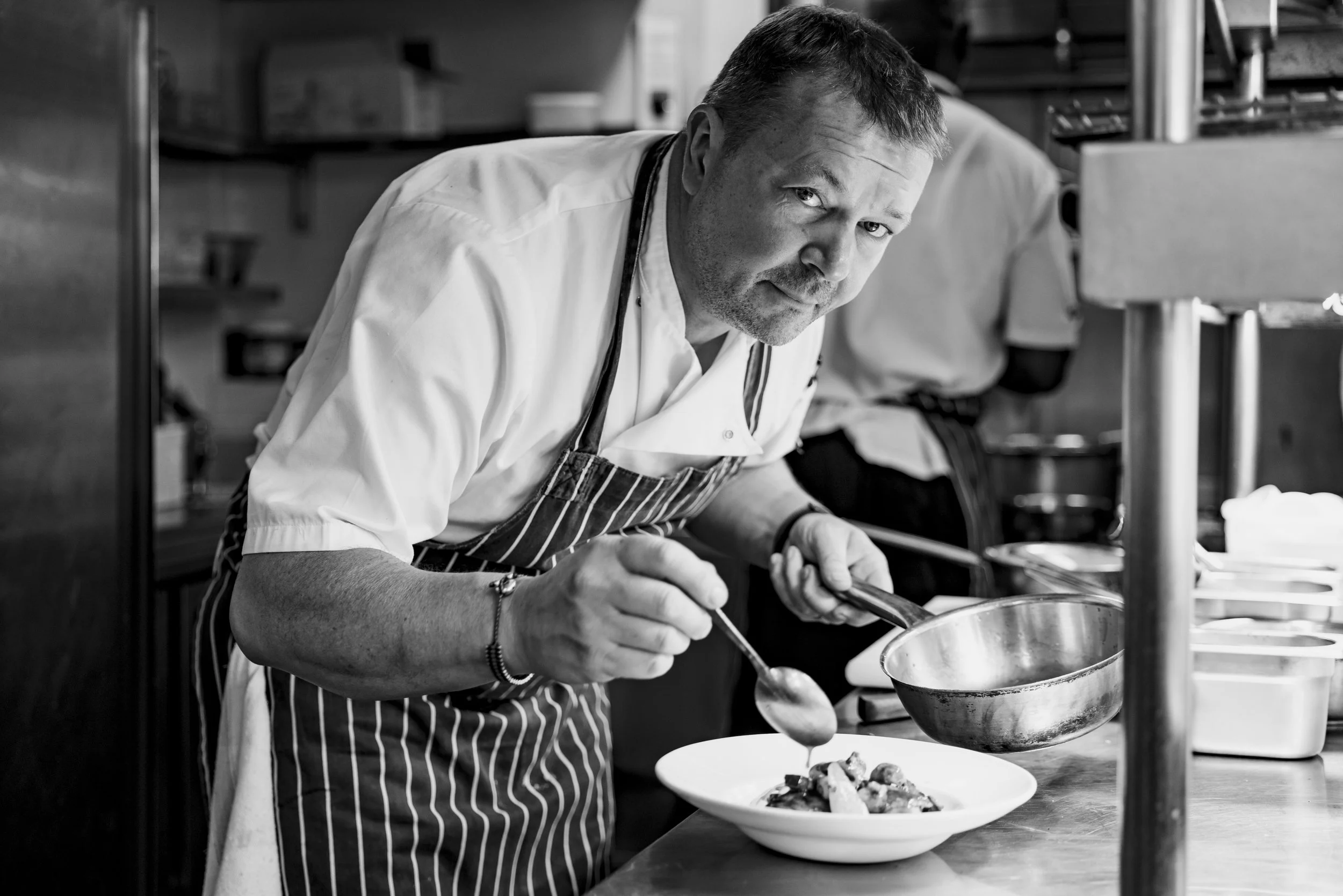 A male chef in a striped apron and white shirt plating food in a professional kitchen, looking at the camera.