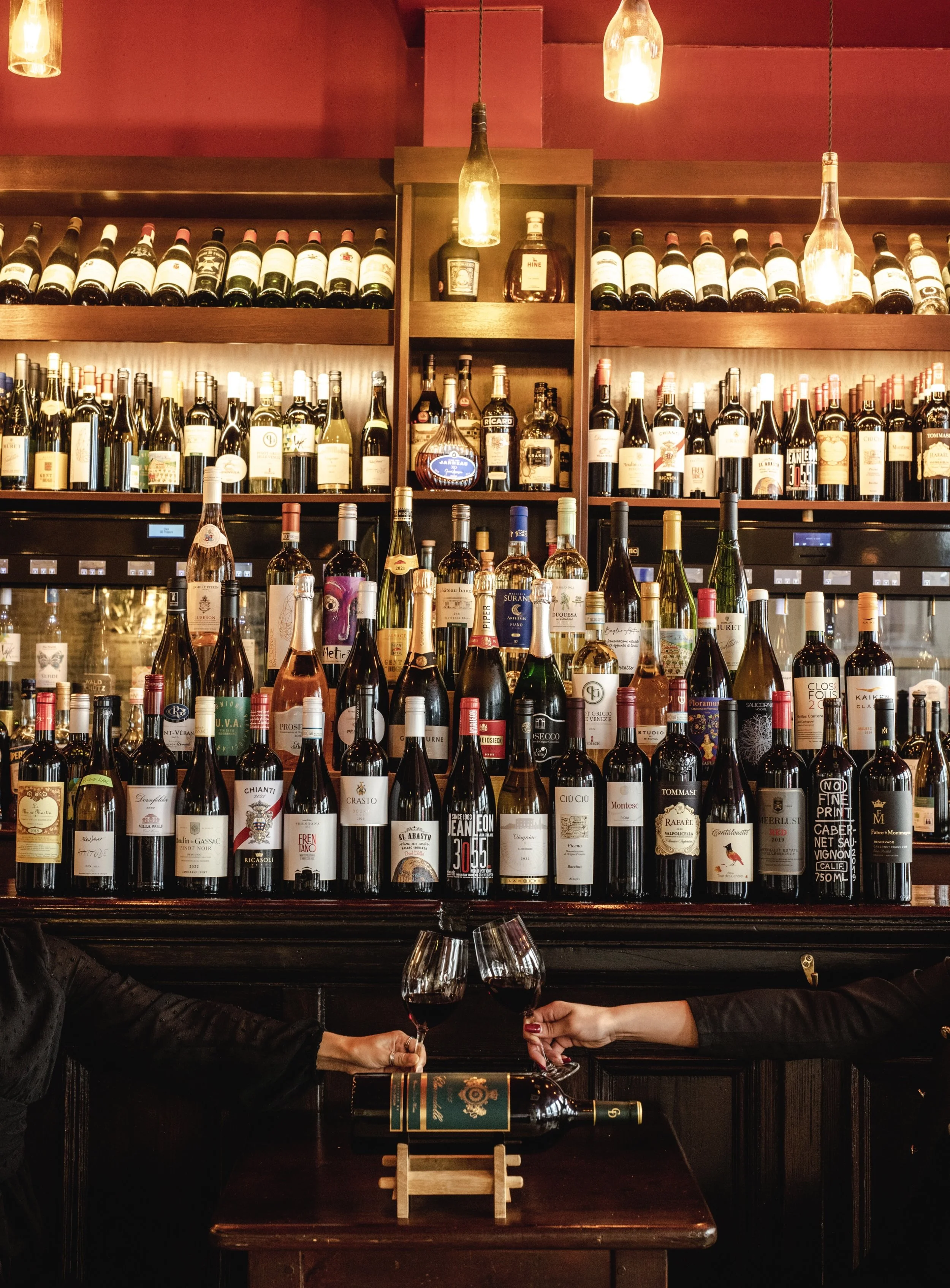 Two people clinking glasses of red wine in front of a bar filled with wine bottles and liquor bottles, with warm lighting overhead.